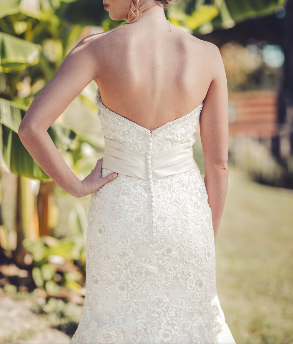 Back view of a bride in a strapless white lace wedding dress with pearl buttons and a satin sash, standing outdoors among green foliage.