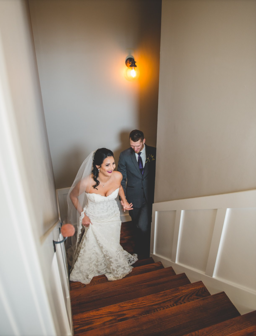 A newlywed couple walking up stairs, with the bride in a white wedding dress and veil, and the groom in a suit, smiling and holding hands.