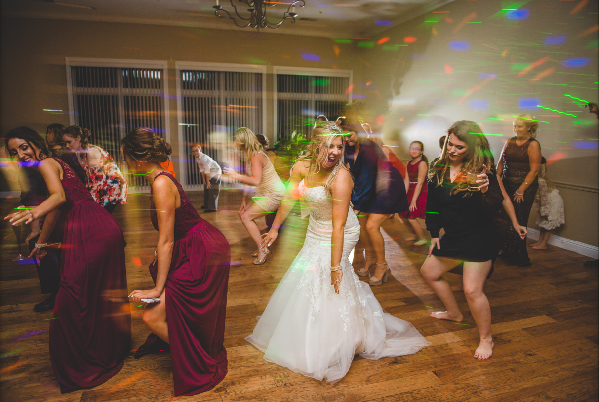 People dancing at a party with colorful lights, including a woman in a white wedding dress and others in formal attire.