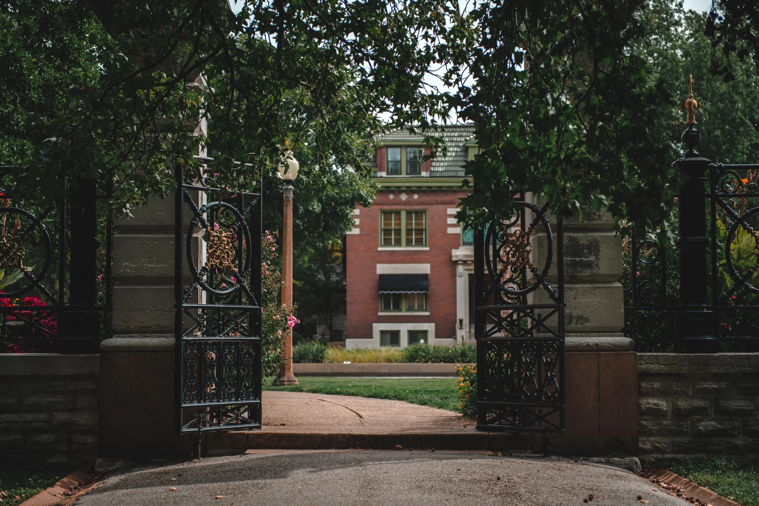 Open wrought iron gate leading to a brick house with trees and bushes in the front yard.