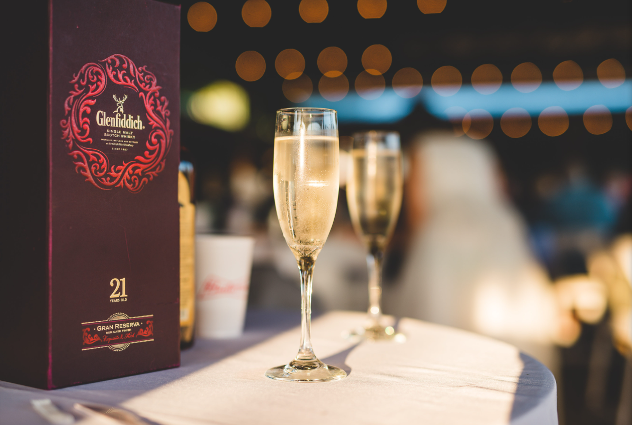 A bottle of Glenfiddich single malt Scotch whisky and two glasses of champagne on a table at a dimly lit event or restaurant.