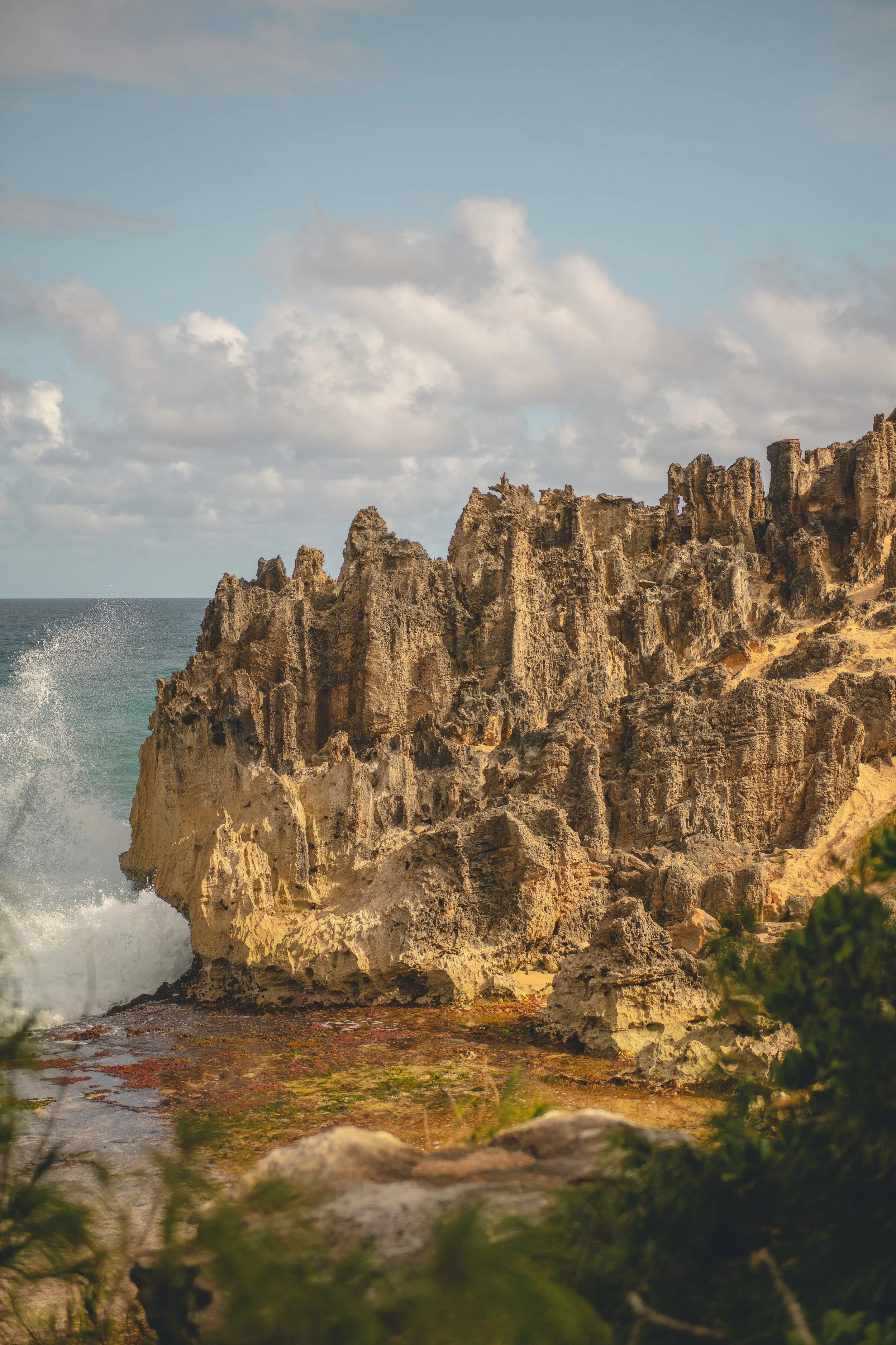 Coastal scene with rugged sandy-colored rocks and cliffs by the ocean under a partly cloudy sky.