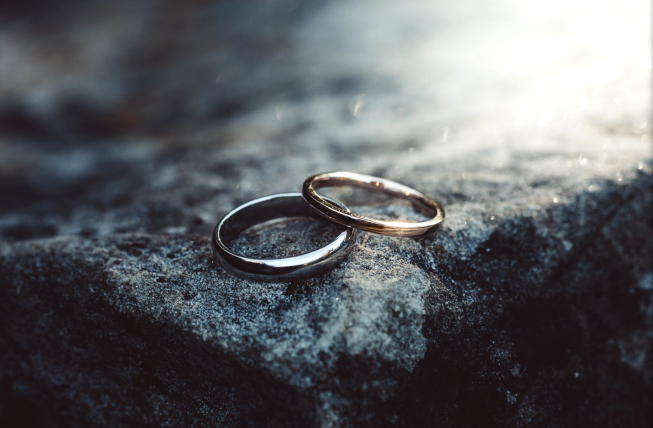 Two wedding bands, one silver and one gold, resting on a textured rock surface with sunlight shining from the right.