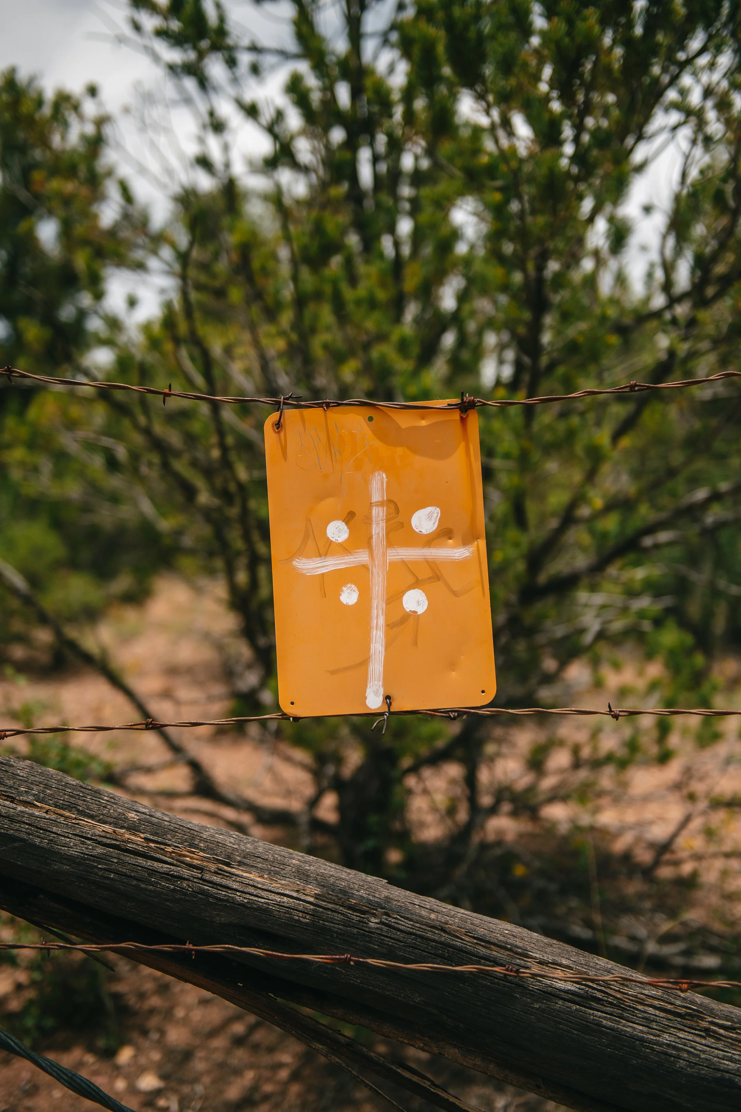 Orange metal sign with a white cross and four dots, hanging on a barbed wire fence with a tree in the background.