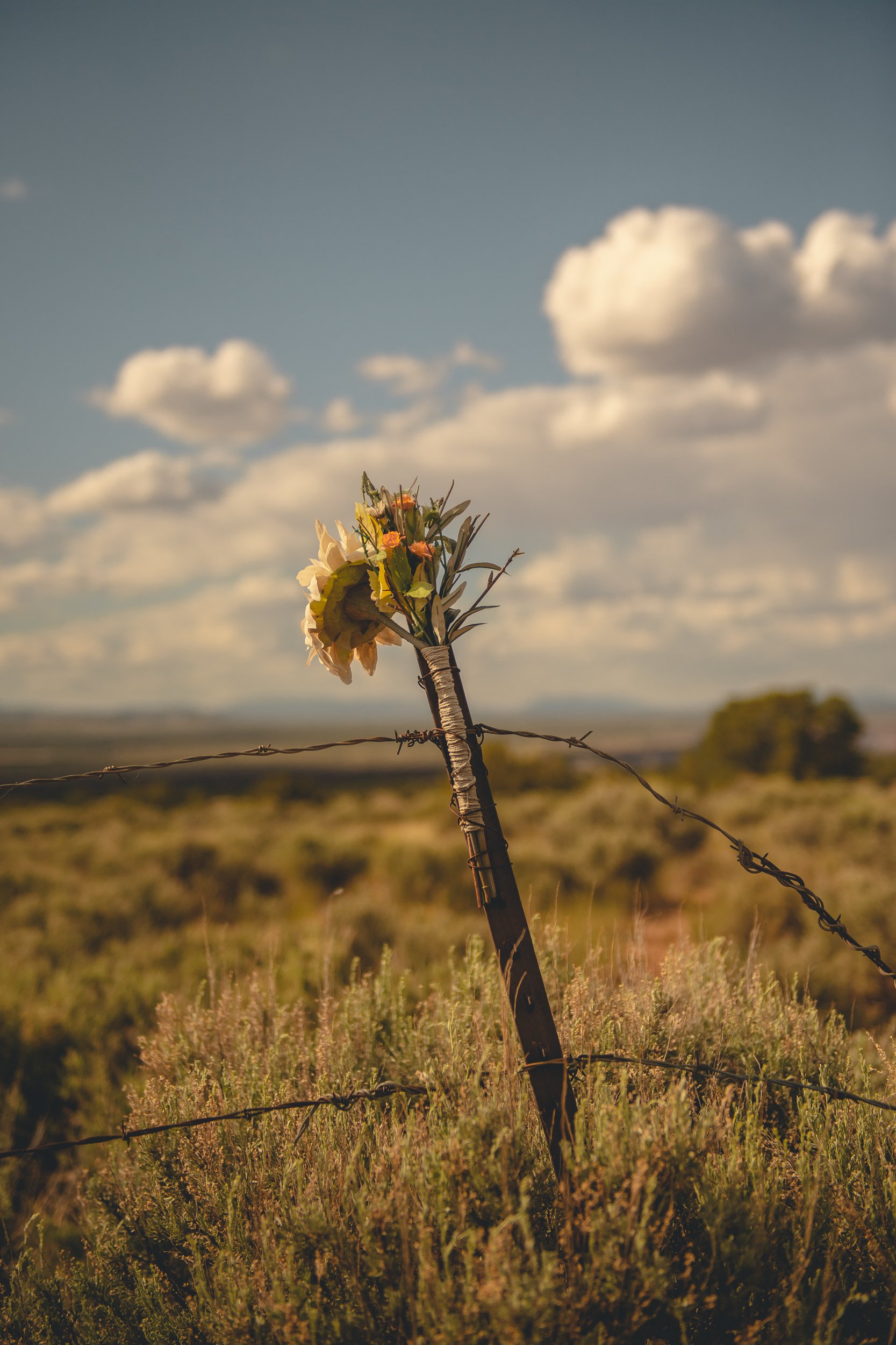 A wooden fence post wrapped with barbed wire has a bouquet of colorful artificial flowers attached to it, set in a rural landscape with open fields and a partly cloudy sky.