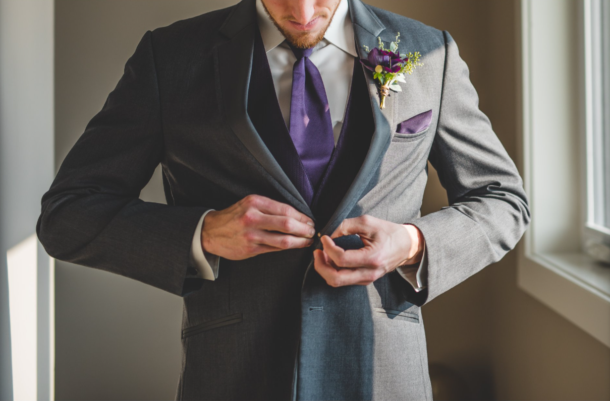 Man in a gray suit with purple tie and boutonniere fastening buttons on his jacket.