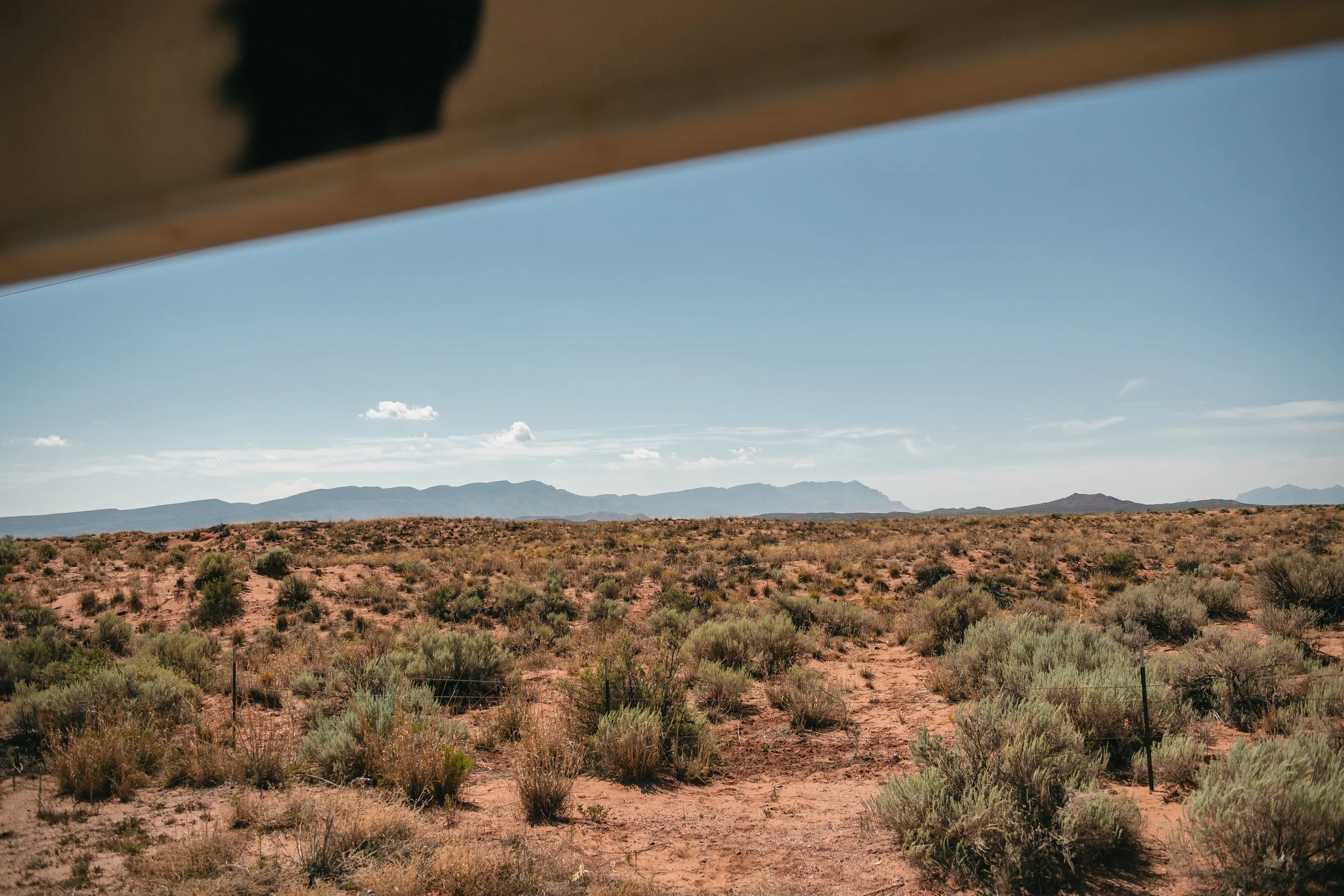 Desert landscape with sparse bushes, mountains in the distance, under a blue sky with a few clouds, seen from beneath a structure.