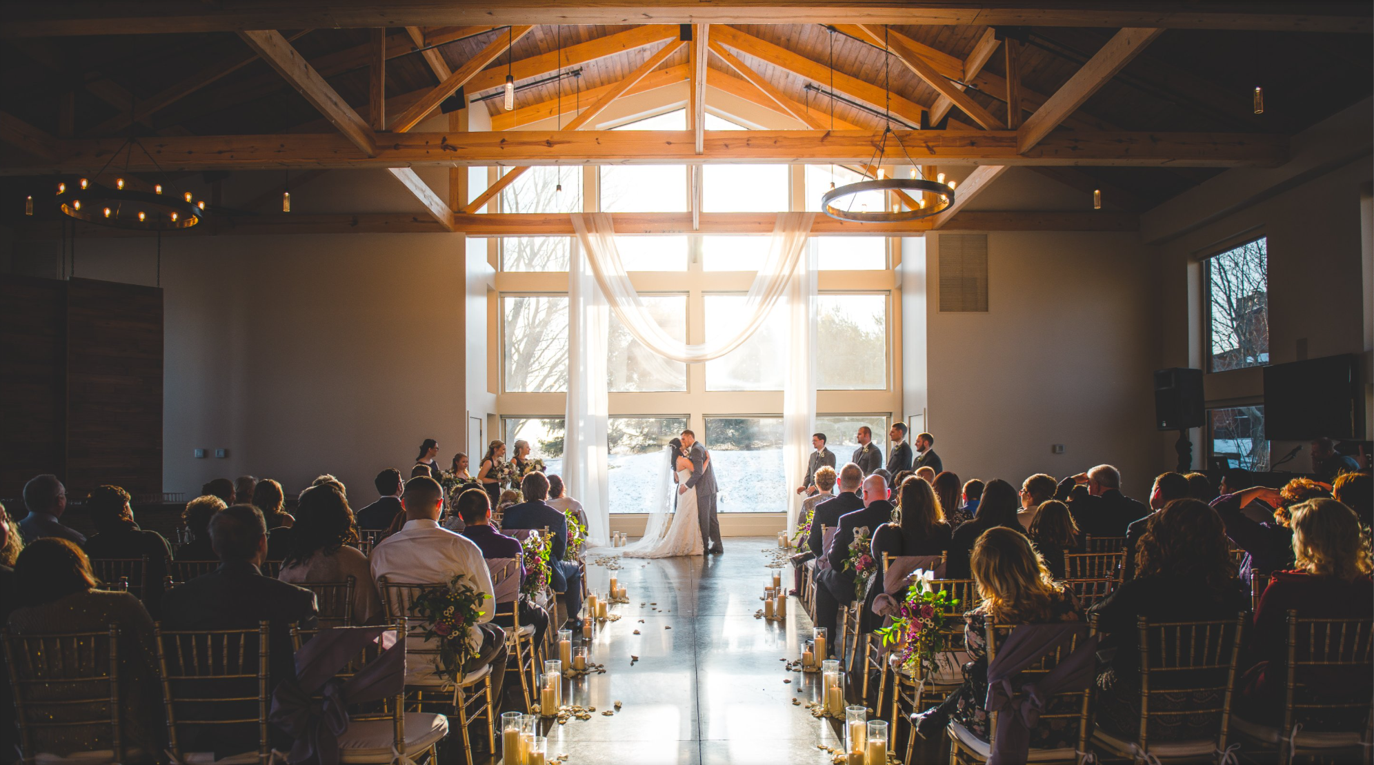 A wedding ceremony taking place indoors with a bride and groom kissing at the altar, surrounded by guests seated in rows, with large windows bringing in natural light and a winter landscape outside.
