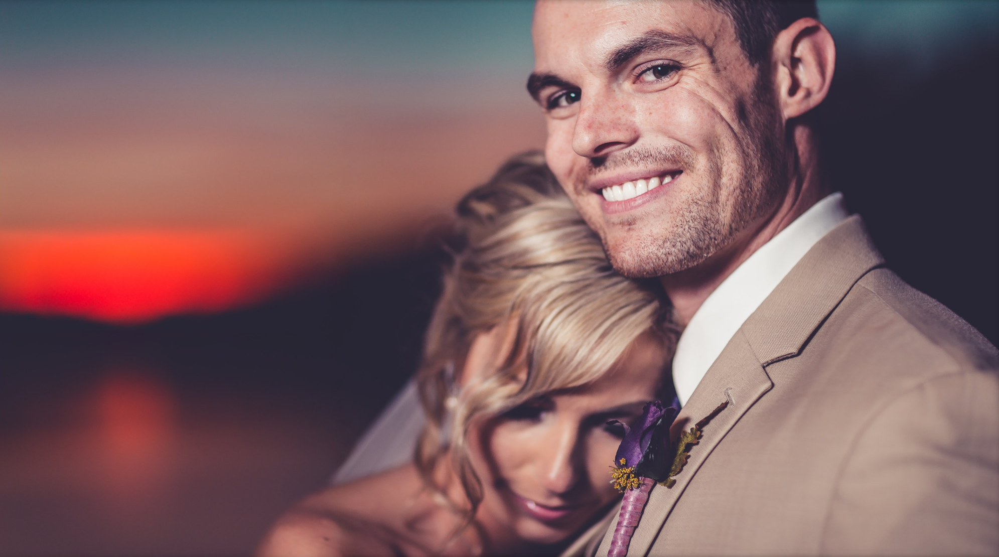 A man with a big smile and a woman with blonde hair hugging each other, outdoors at sunset, dressed in formal wedding attire.