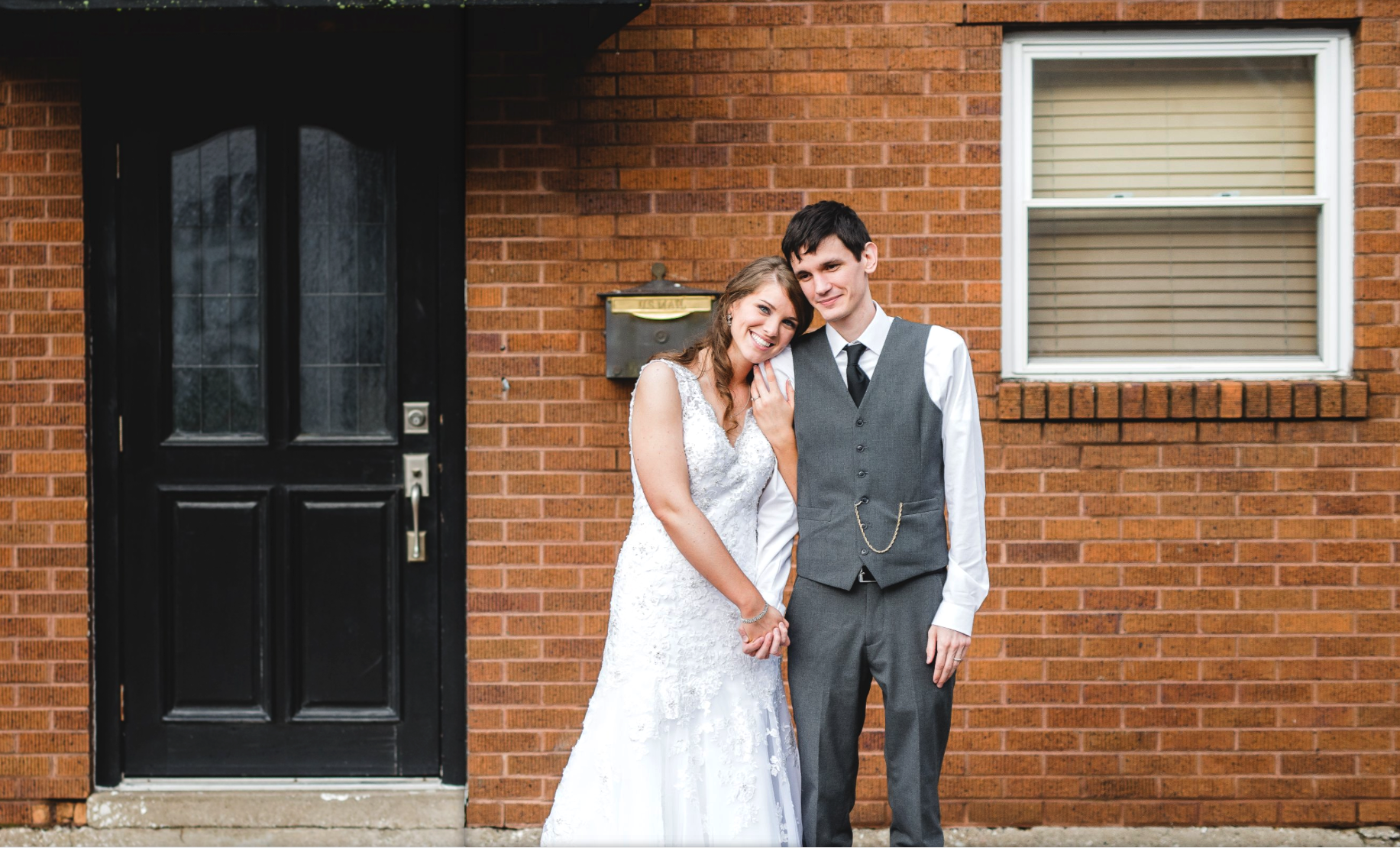 A bride and groom standing outside a brick house, holding hands and smiling at the camera.