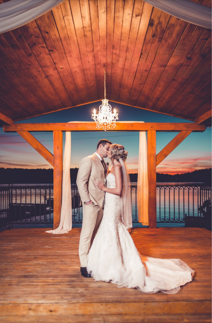 A bride and groom share a kiss under a lit chandelier on a wooden deck overlooking a river at sunset.