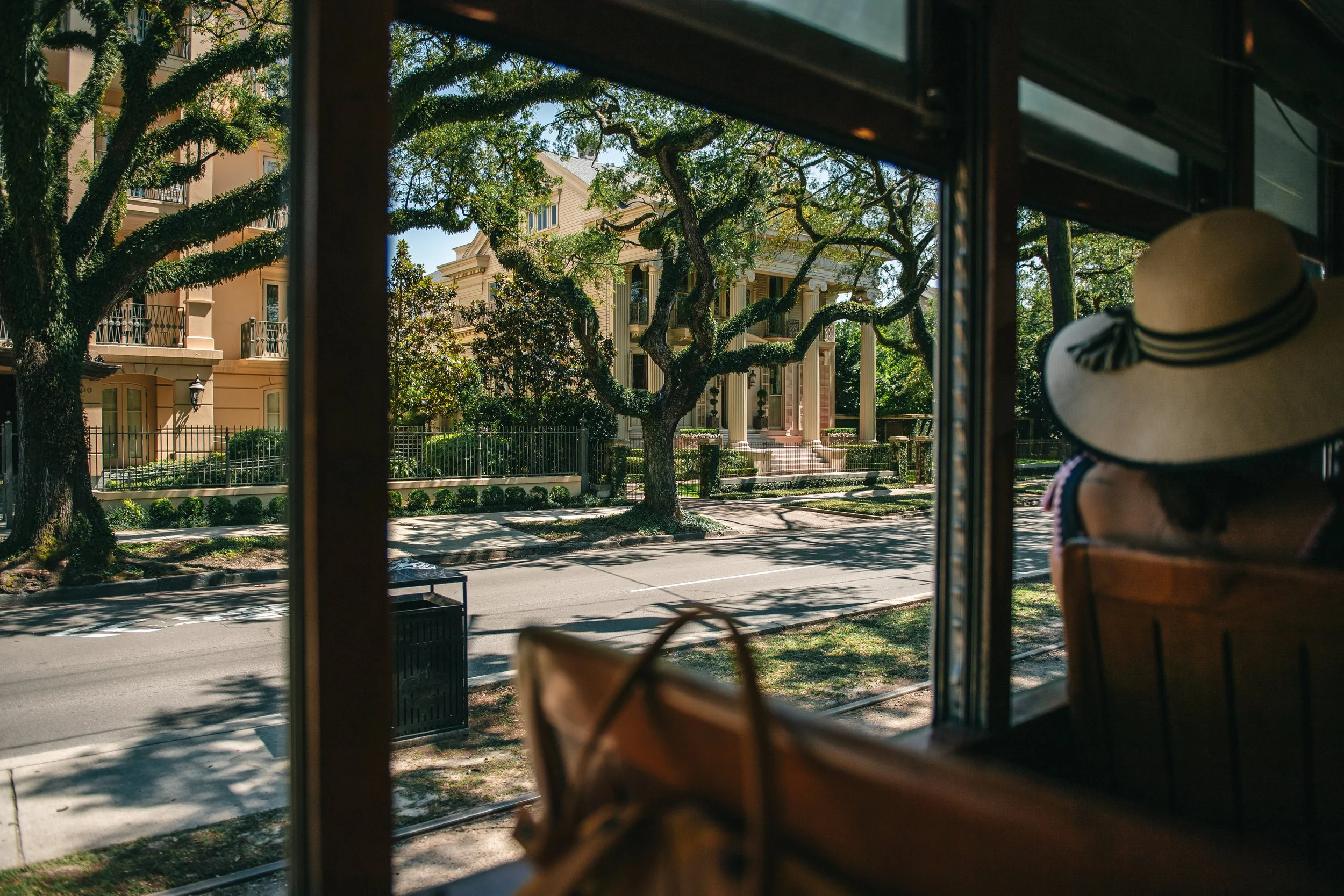 View of a tree-lined street outside a historic house with columns, seen from inside a cafe or shop through a window, with a woman wearing a large sun hat sitting inside.