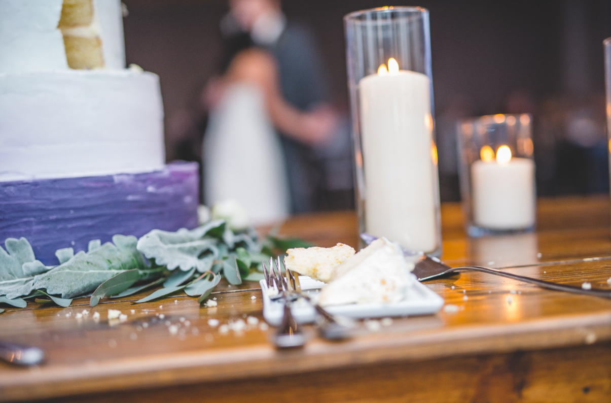 A wedding cake on a table with two candles in glass holders and a plate with a slice of cake, forks, and crumbs.