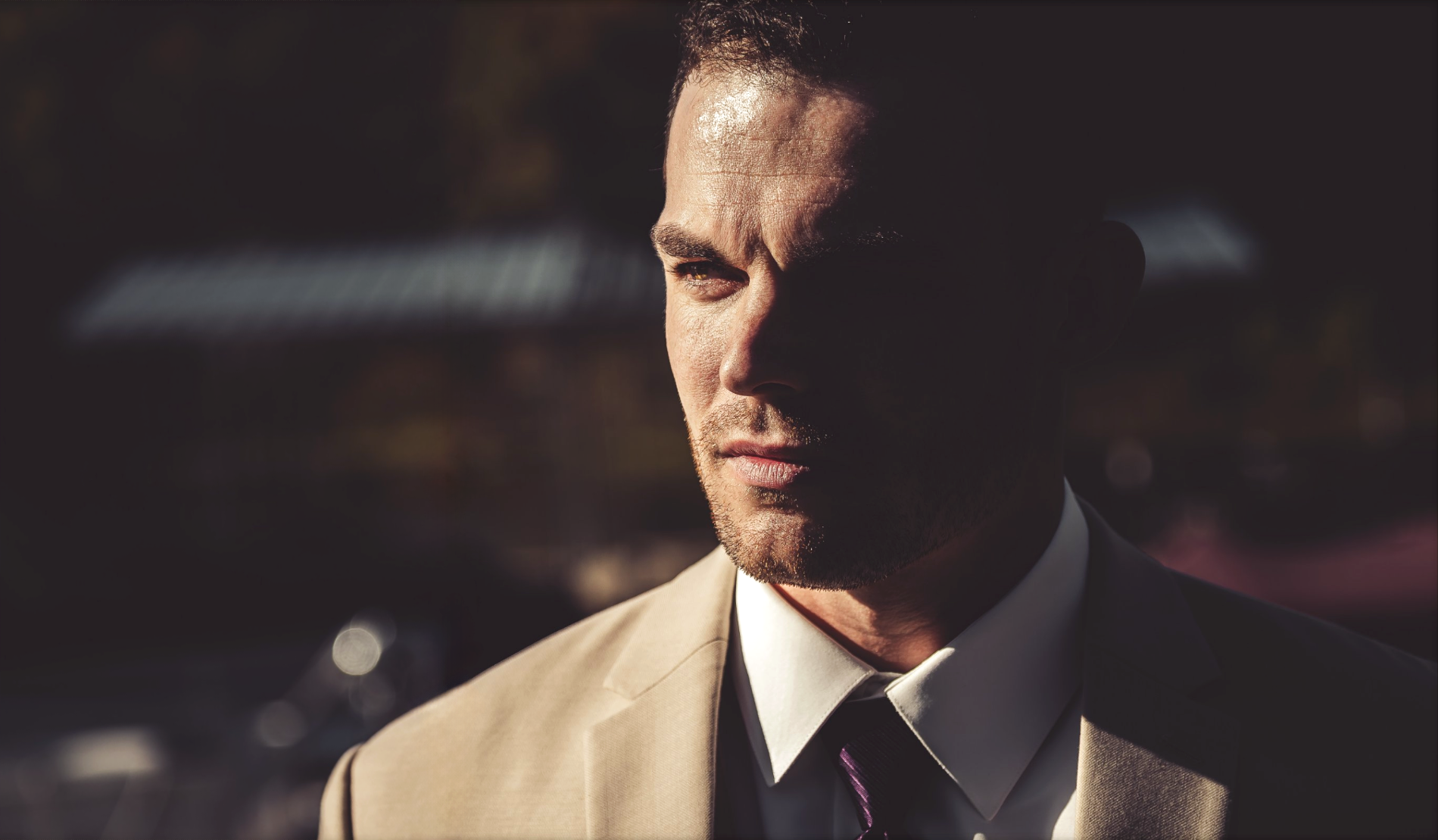 A man dressed in a beige suit and white shirt, wearing a black tie, gazes thoughtfully into the distance outdoor during late afternoon or evening, with sunlight casting shadows on his face.