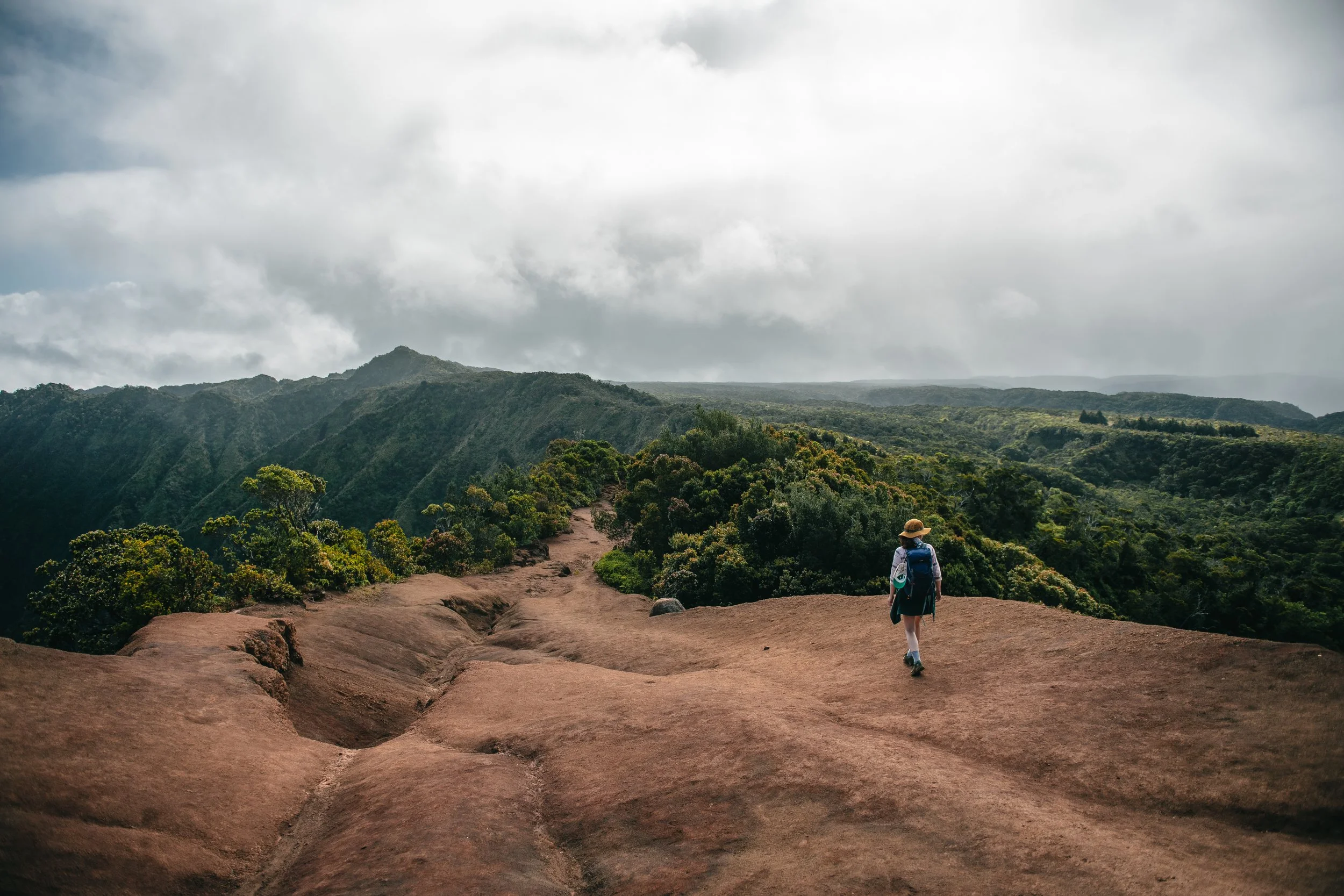Person hiking on a rocky trail through lush green mountains under a cloudy sky.
