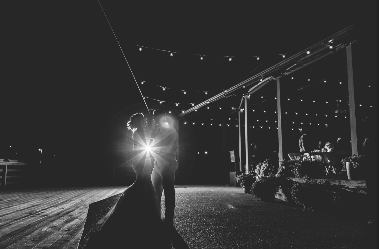 A black and white photo of a couple dancing closely under string lights at night, with the sun or a bright light source creating a starburst effect between them.