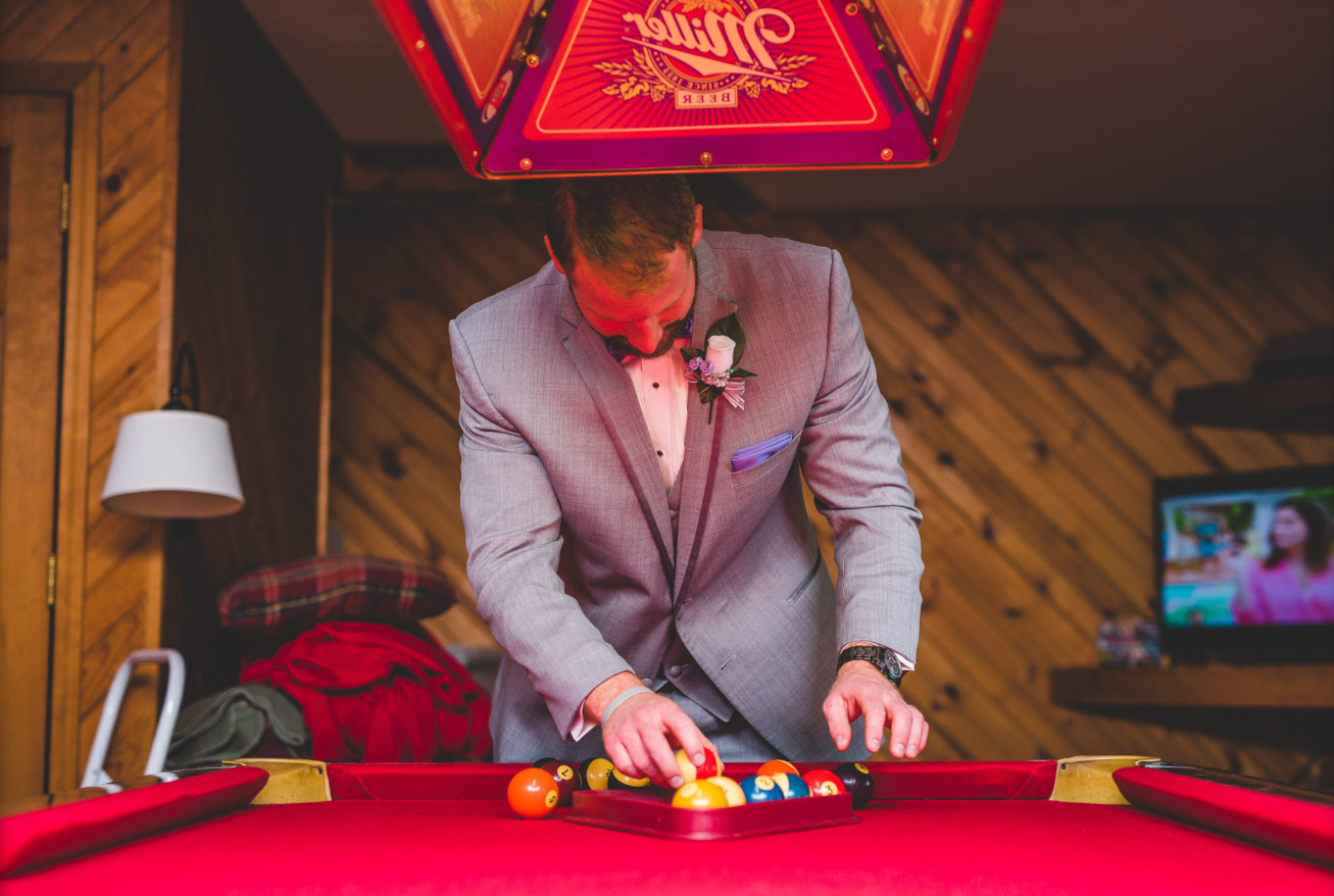 A man in a grey suit with a flower boutonniere playing pool in a wood-paneled room with a red table and a television in the background.