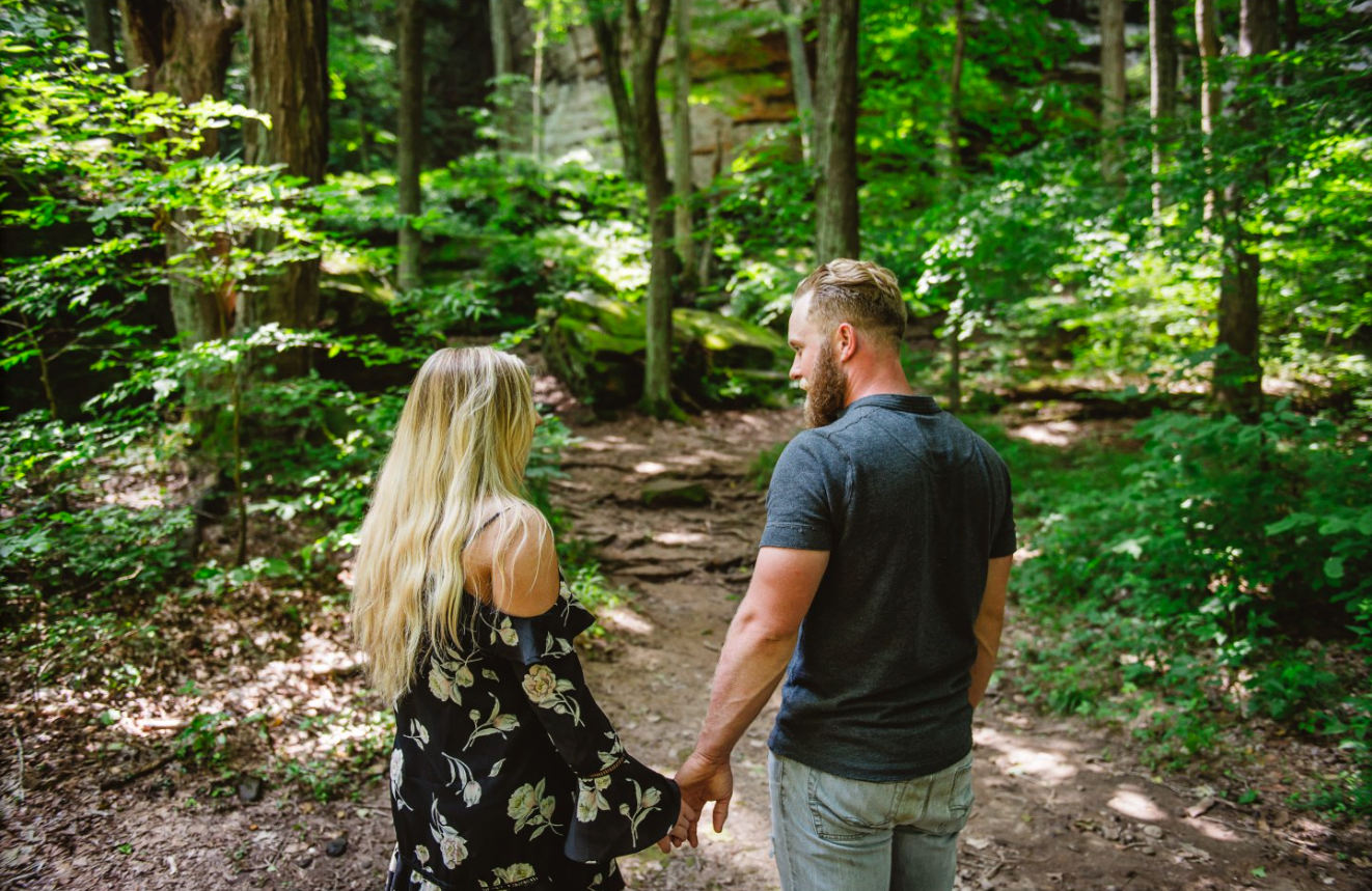 A man and a woman holding hands walking on a dirt trail in a green forest.