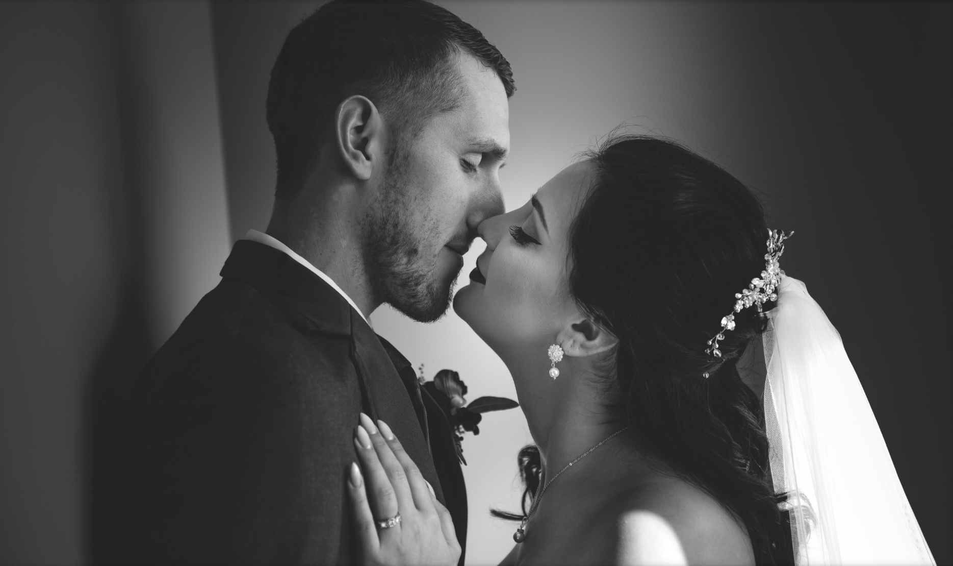 Black and white photo of a bride and groom about to kiss, with the bride wearing a tiara, earrings, and a wedding dress, and the groom in a suit, holding each other closely.