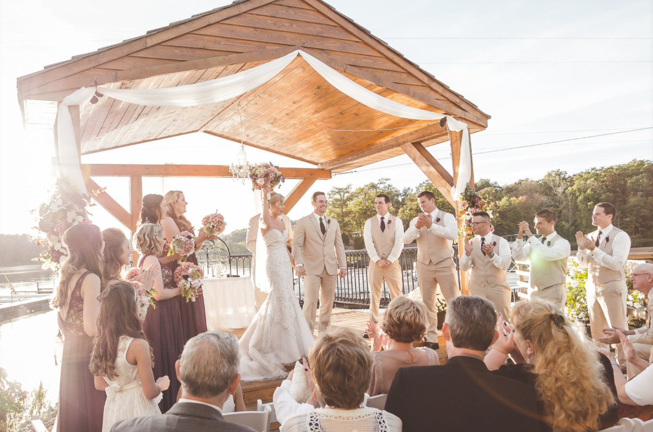 Outdoor wedding ceremony on a wooden stage with a bride, groom, bridesmaids, groomsmen, and guests, set against a lakeside backdrop at sunset.