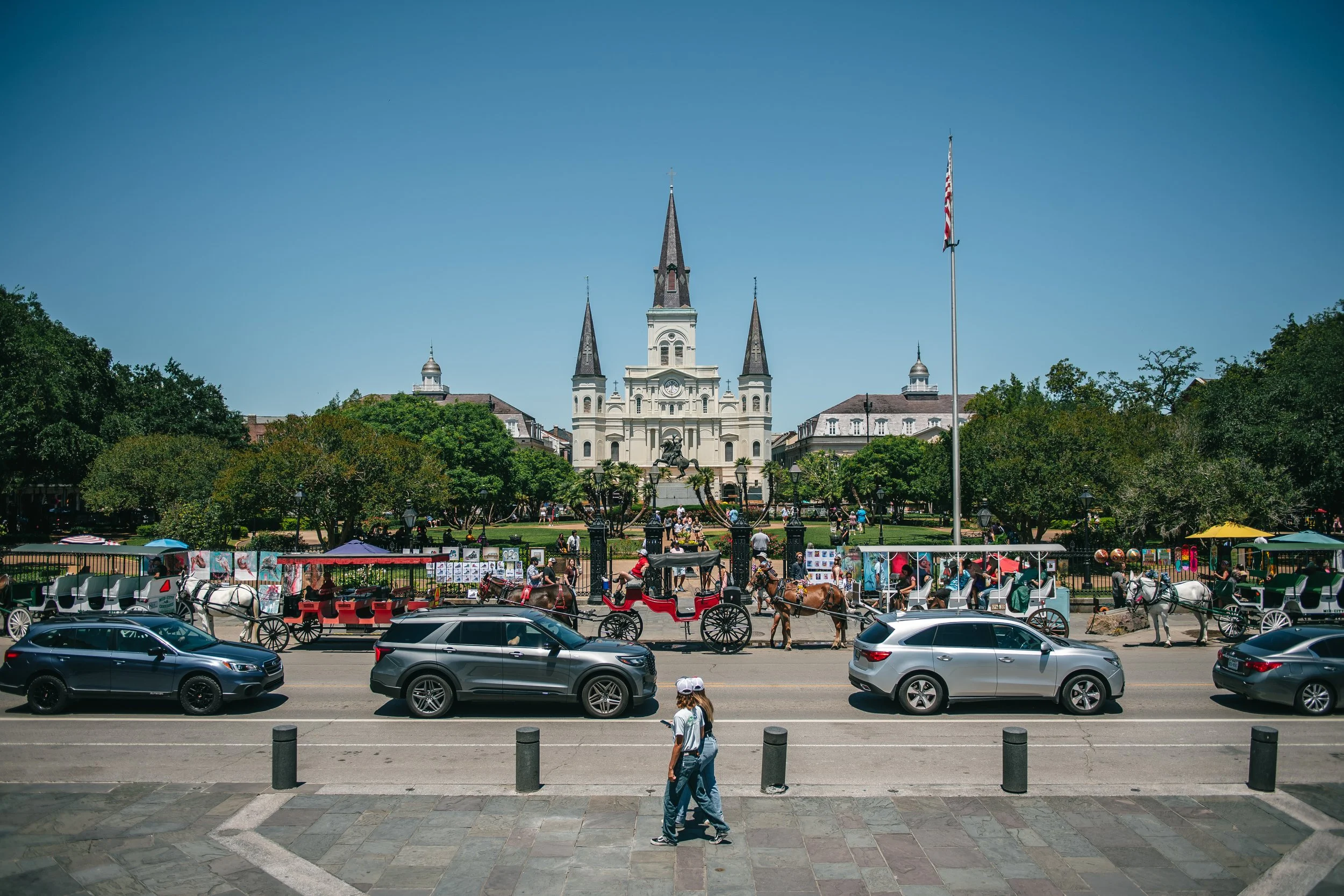 Jackson Square with Saint Louis Cathedral in New Orleans. Front view of the square with people walking and cars on the street, horse-drawn carriages parked along the sidewalk, and a bright blue sky.