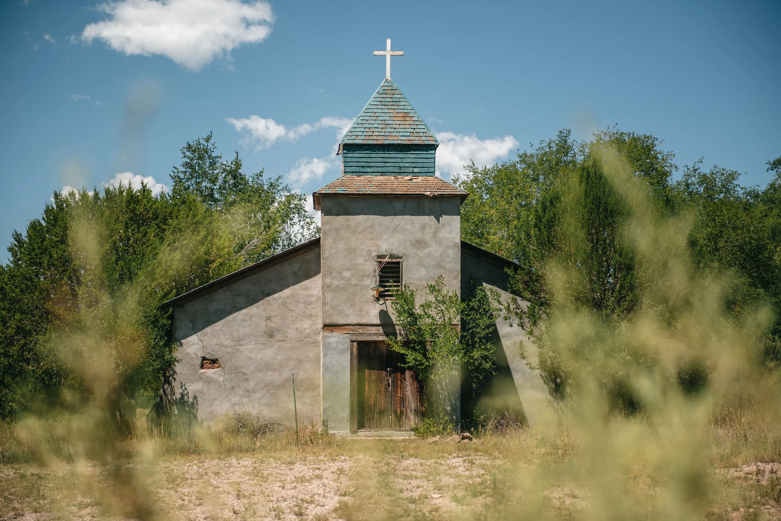 Old small church building with a cross on top, surrounded by trees and grass, under a blue sky with clouds.