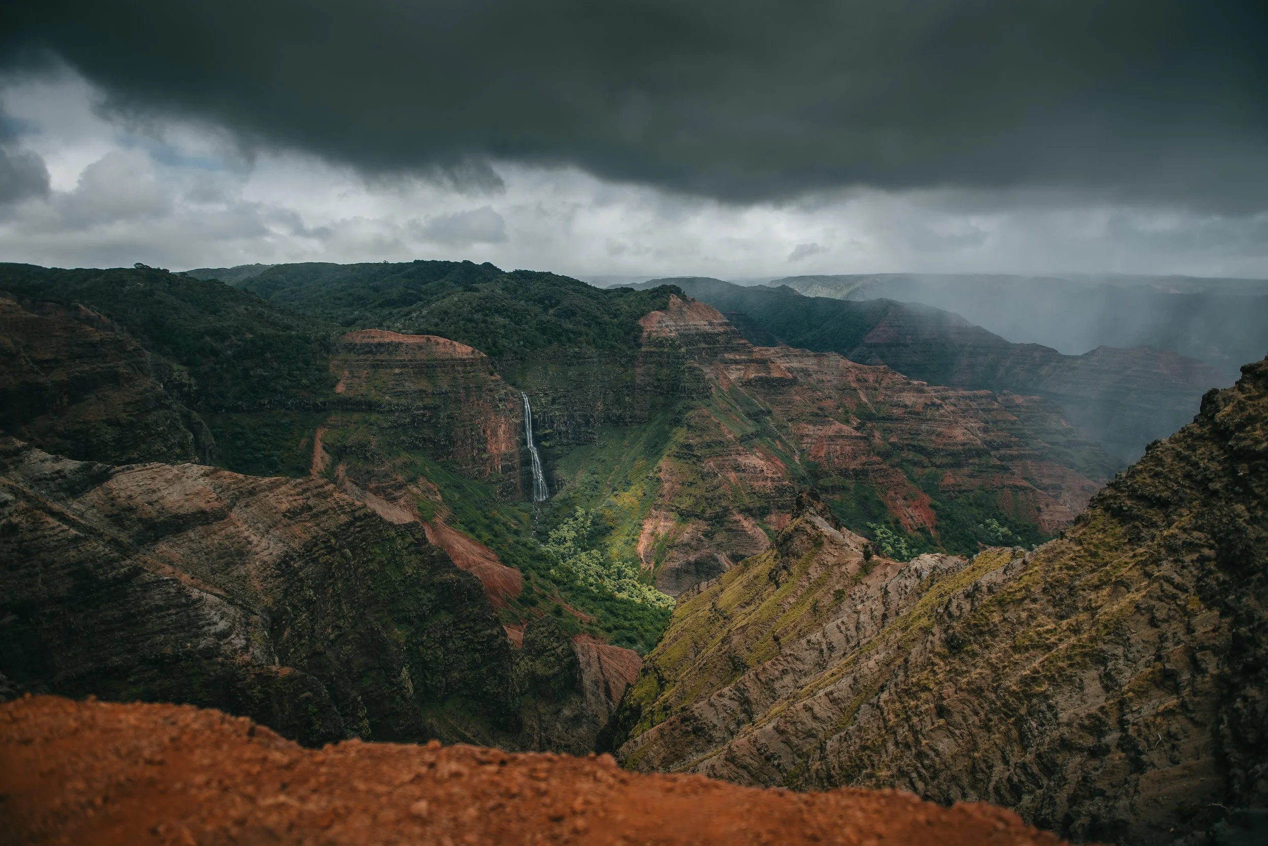 Overcast skies above the lush green and reddish-brown cliffs of a canyon, featuring a waterfall cascading down the side.