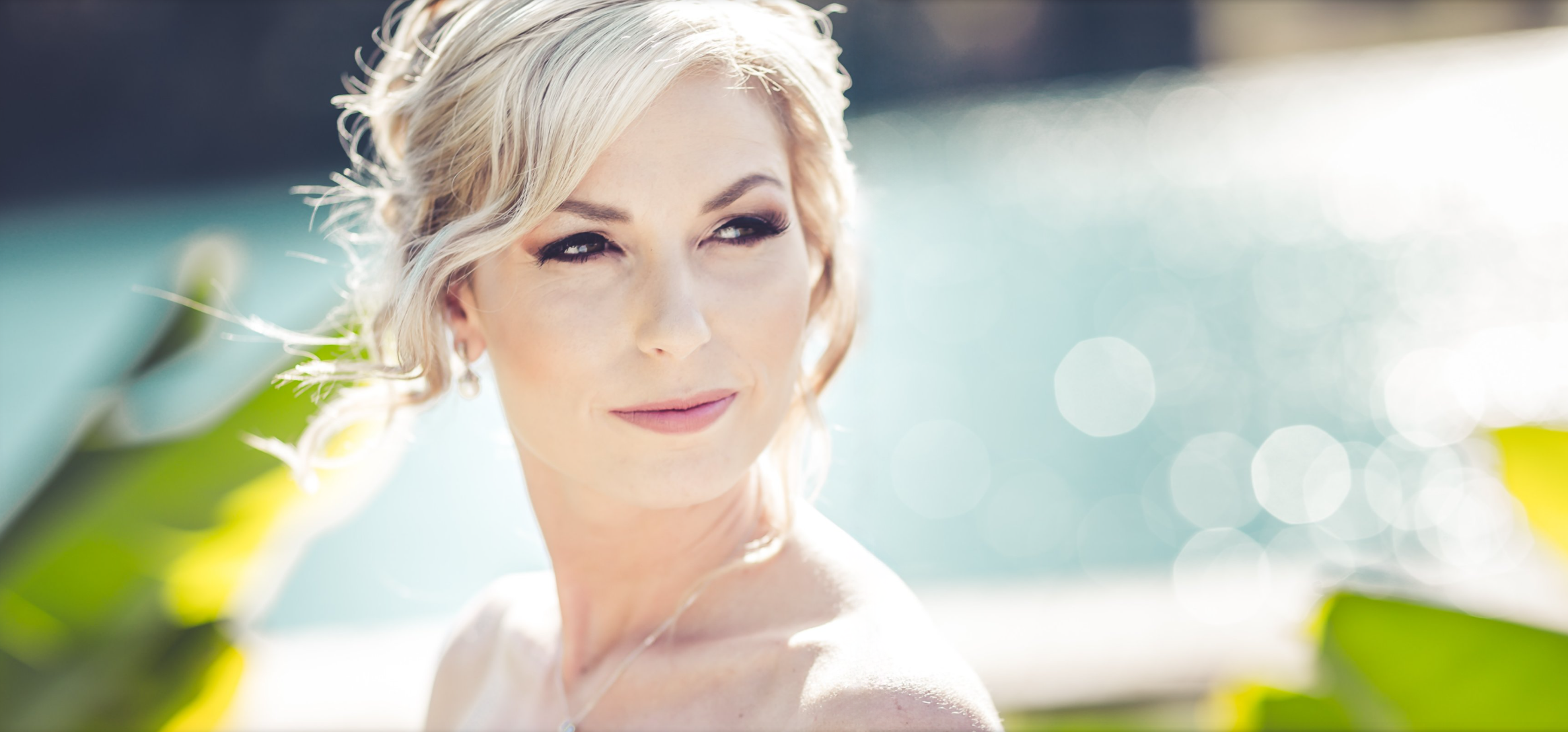 Close-up of a woman with blonde hair outdoors near water, smiling softly and looking to the side with sunlight lighting her face.