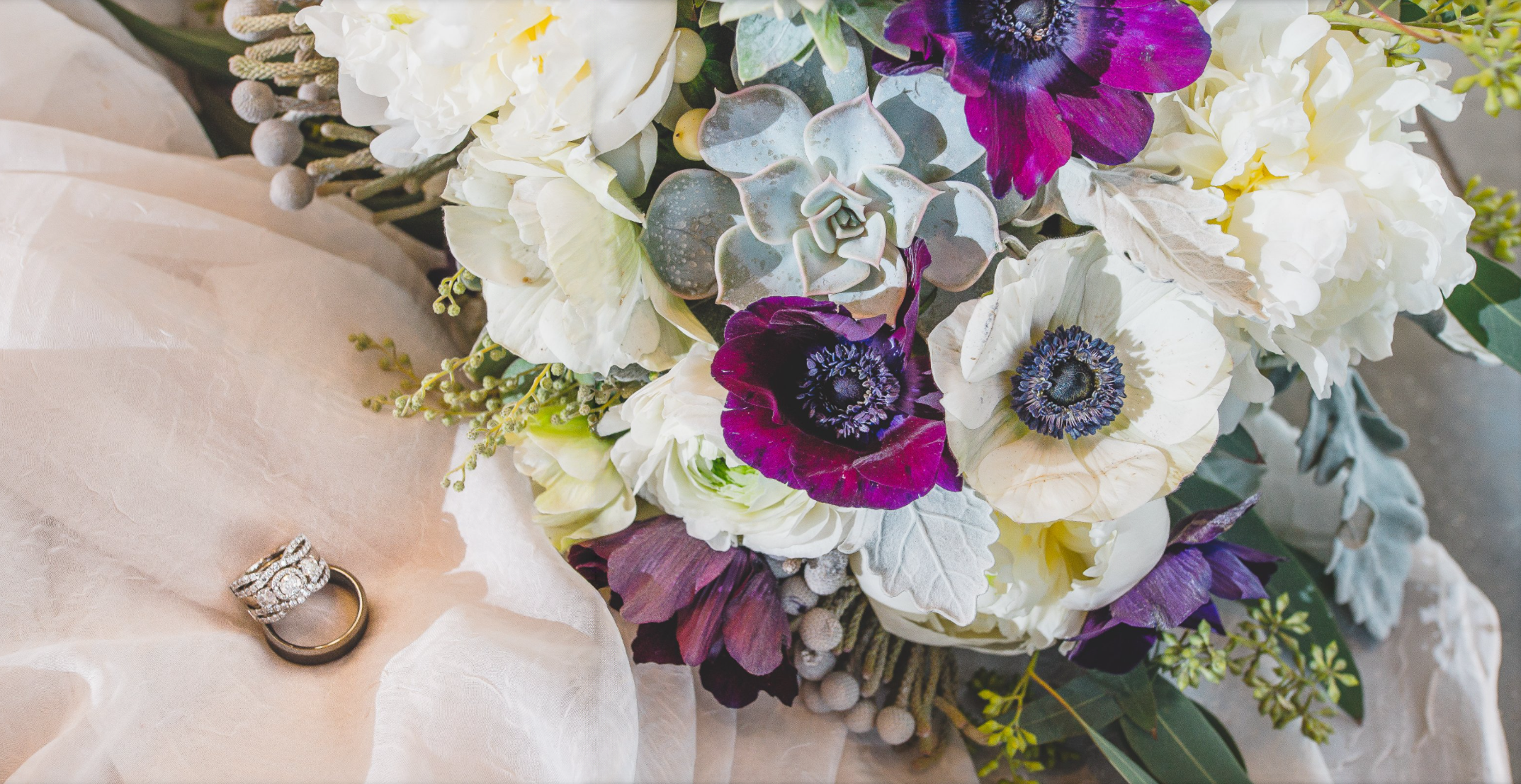 A bouquet of white, purple, and gray flowers with two rings, one with diamonds, resting on beige fabric.
