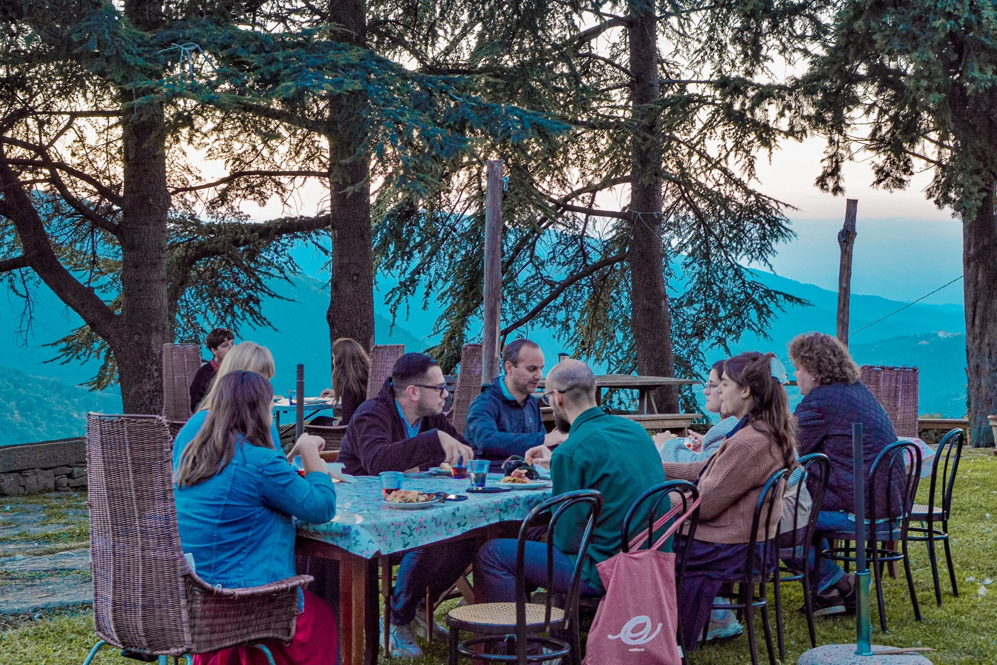 People dining outdoors at a mountain-side restaurant during the evening, surrounded by tall trees and scenic mountain views in the background.