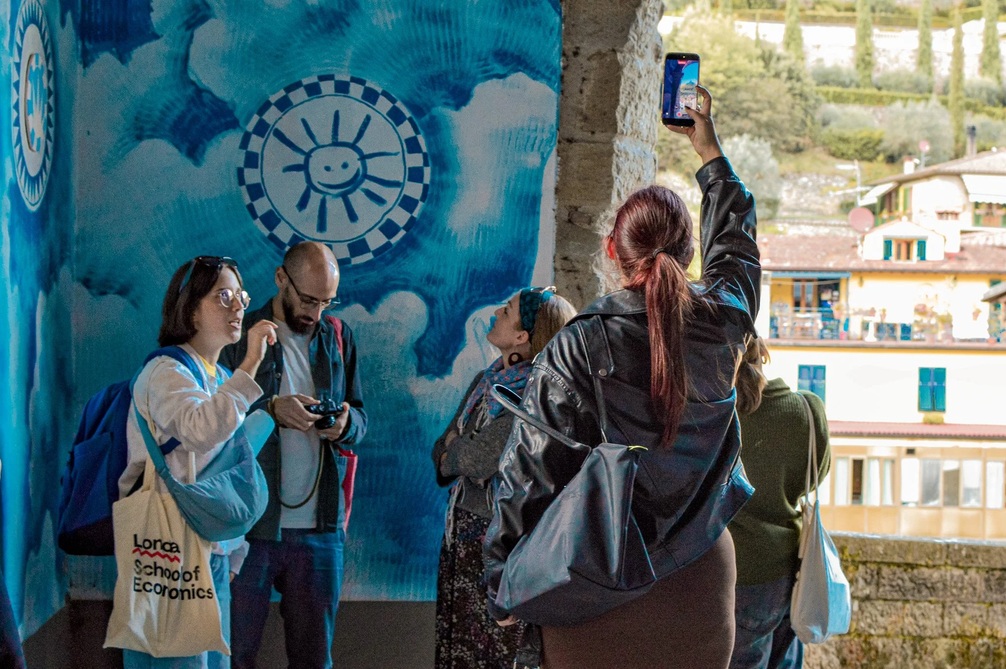 Group of people standing near a blue wall with a smiling sun drawing, some using phones or cameras, outside with buildings and trees in the background.
