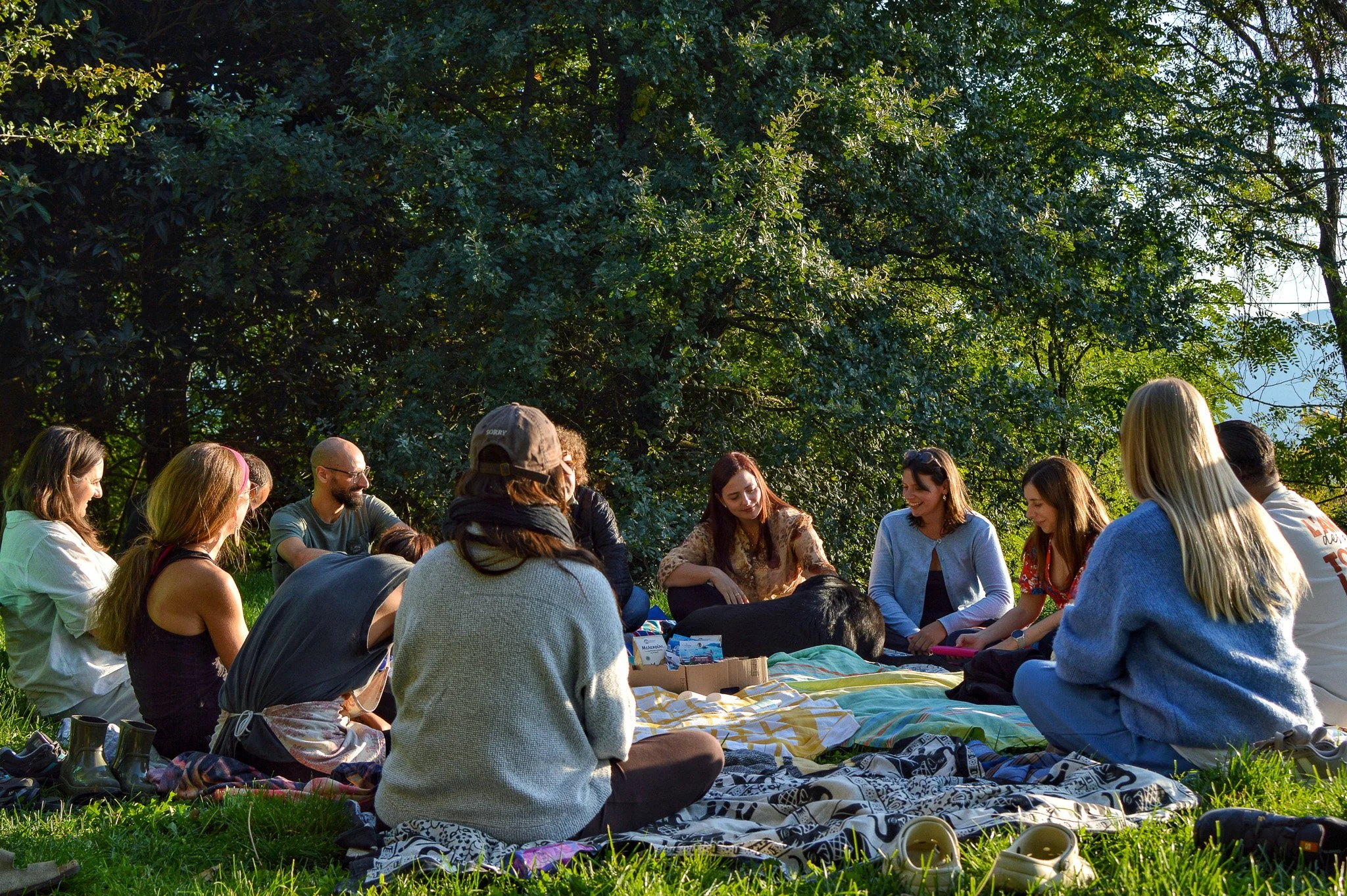 A group of people sitting closely together outdoors on grass, under trees, with some items and shoes spread on a blanket, engaging in conversation or activity.