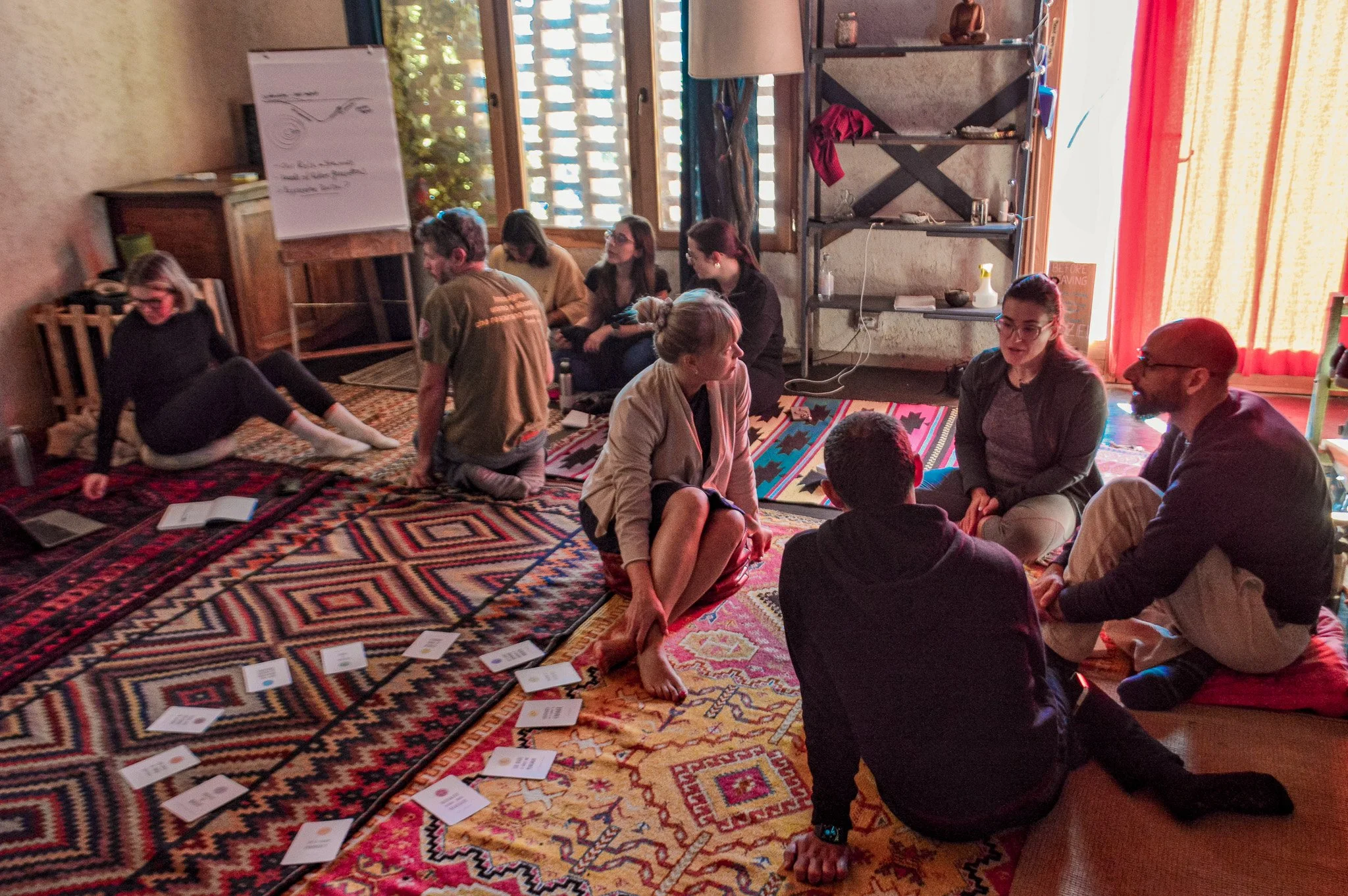 A group of people sitting on colorful patterned rugs in a cozy room, engaged in conversation, with a whiteboard showing a diagram in the background.