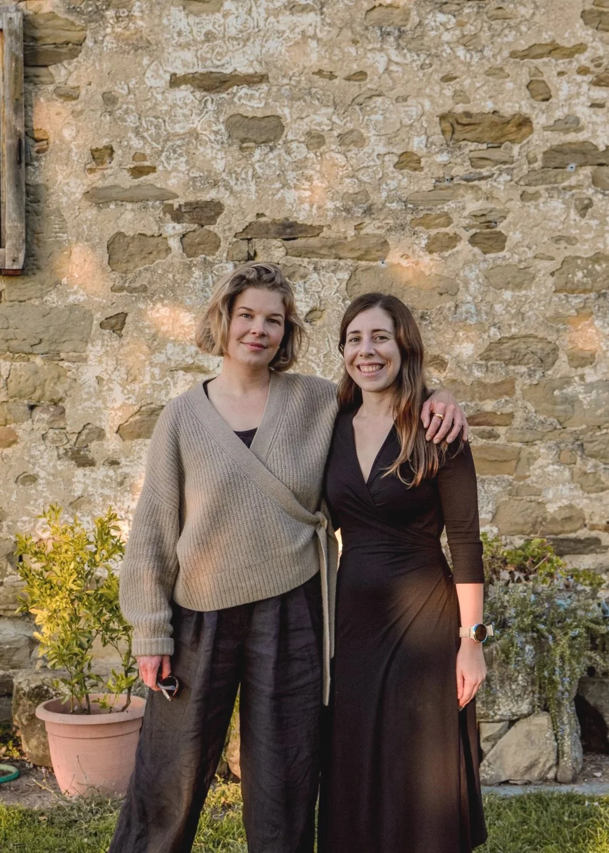 Two women standing outdoors in front of a stone wall, smiling and posing for a photo. One woman has short blonde hair, wearing a beige sweater and black pants. The other has long brown hair, wearing a black dress, with her arm around the first woman.