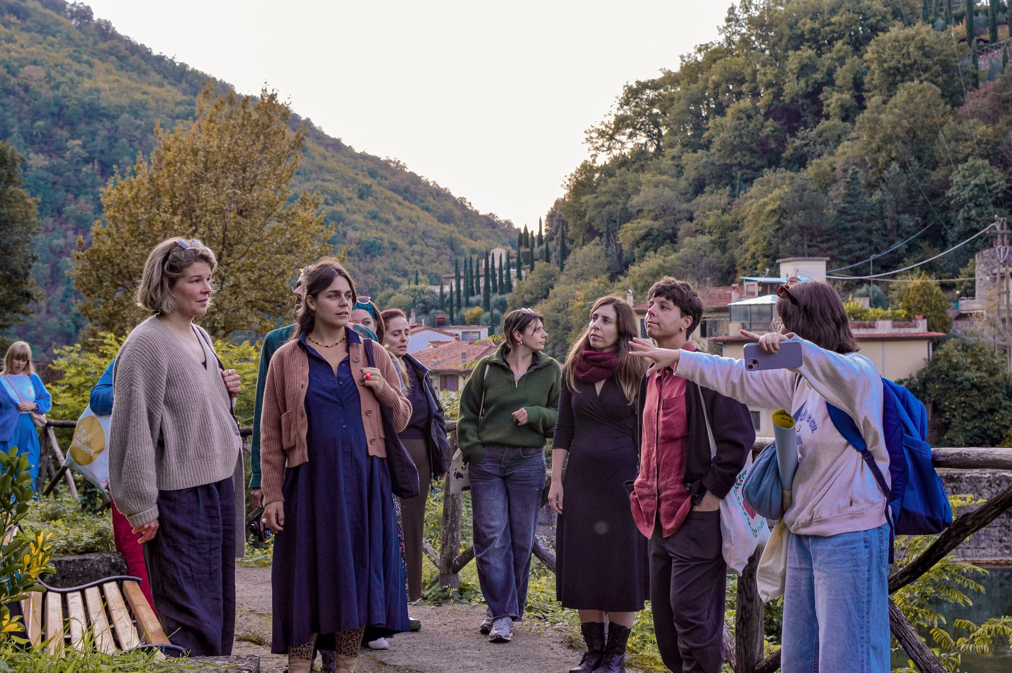 Group of people outdoors in a hilly area with trees and houses, some listening while a woman in a white jacket gestures as she speaks.