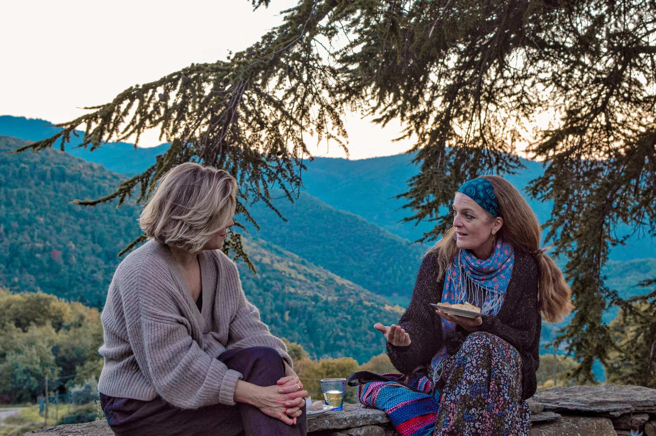 Two women having a conversation outdoors in a mountainous area, one holding a plate with food, under a large tree with mountains in the background during sunset.