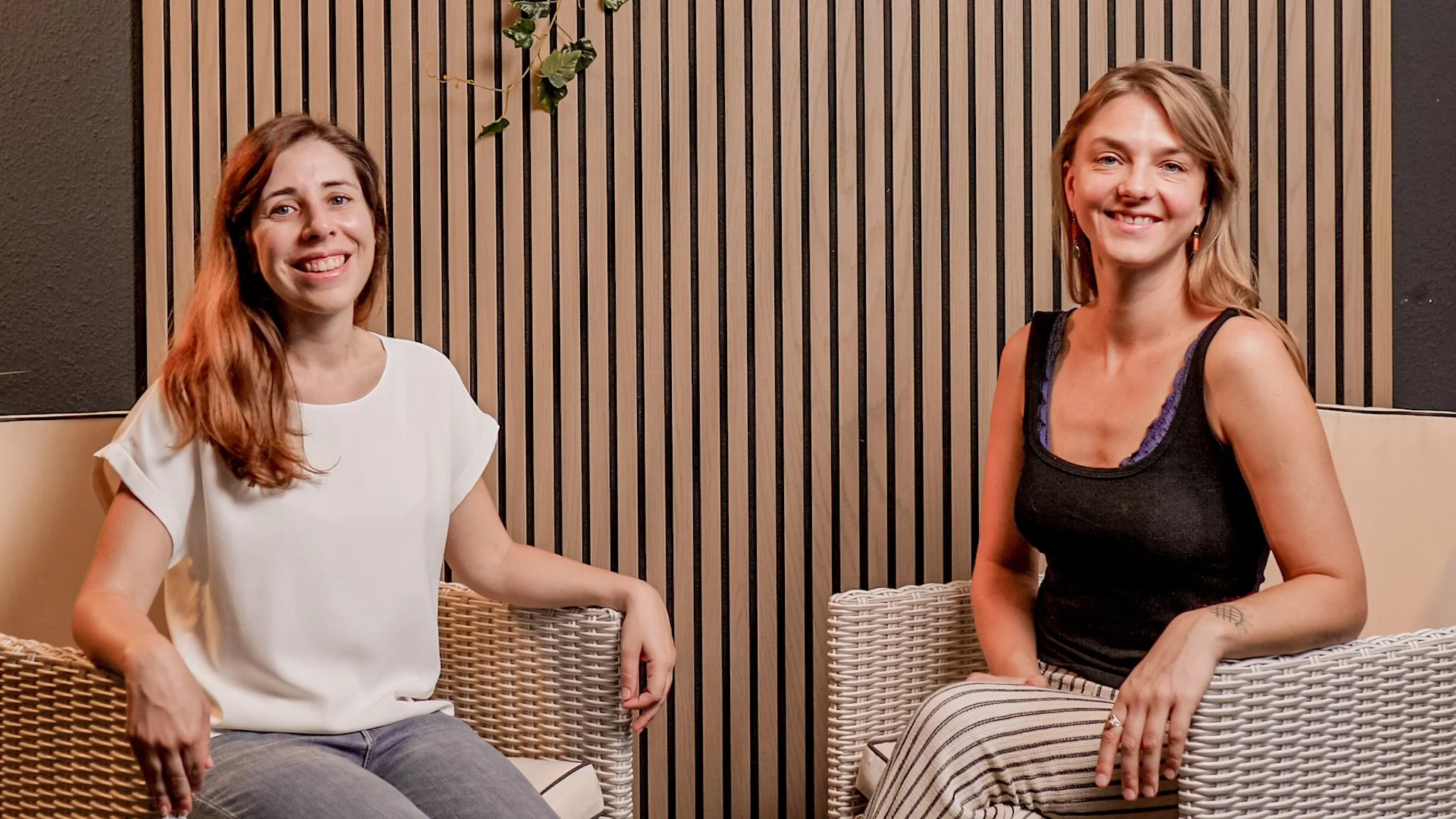 Two women sitting on wicker chairs in a room with wooden panel wall. The woman on the left has long brown hair, wearing a white top, and the woman on the right has shoulder-length blonde hair, wearing a black tank top and striped pants. Both are smiling.