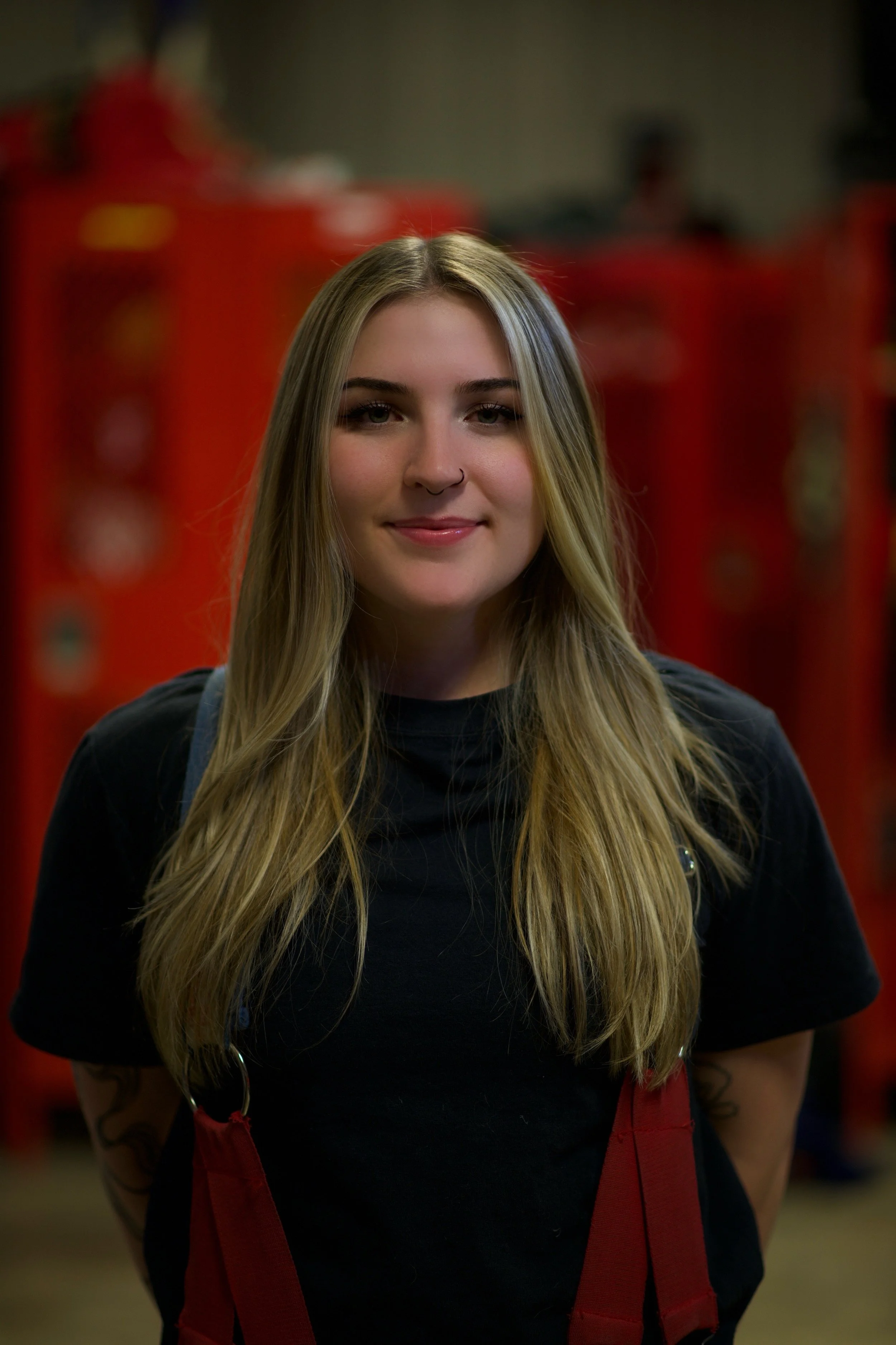 Young woman with long blonde hair, wearing a black shirt and red apron, standing indoors with a blurred red background.