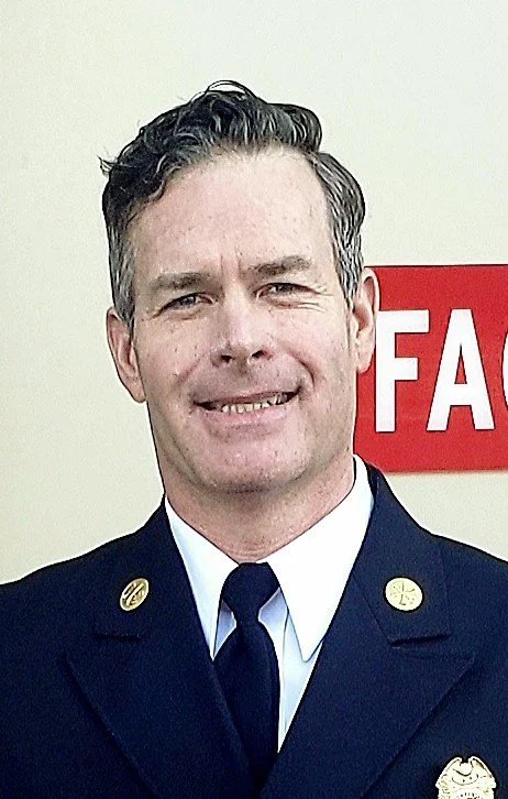 A man in a firefighter uniform smiling in front of a wall with a red and white sign.