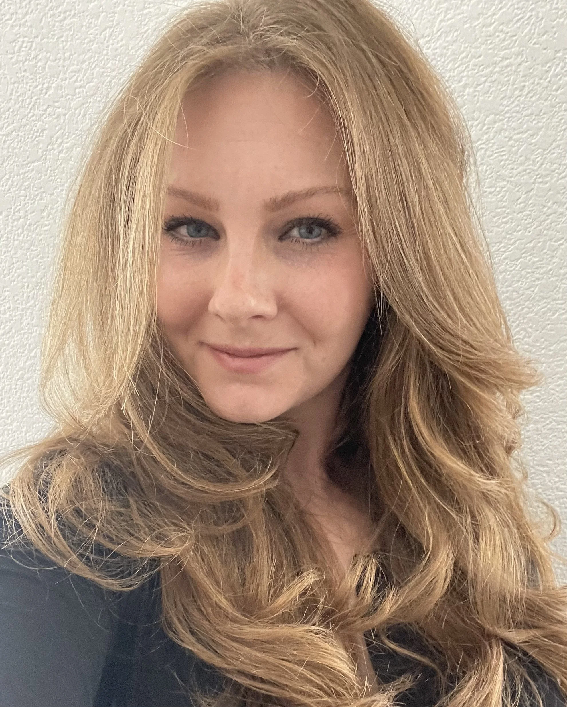 A woman with long, wavy, red hair, blue eyes, and fair skin smiling softly against a textured light-colored wall.