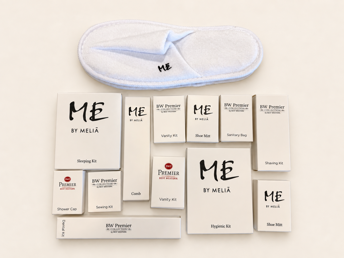 Hotel amenity kits and white slippers arranged on a surface, including sleeping, vanity, shoe mitt, shaving, hygiene, dental, sewing, and shower cap kits.
