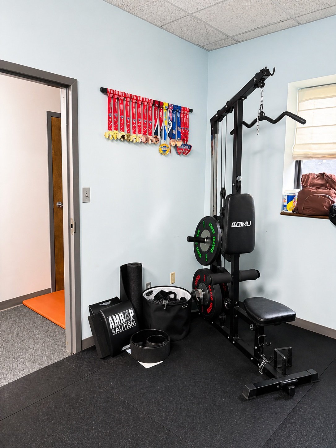 A home gym corner with a black workout machine, a bulletin board with medals, a shelf with water bottles, and a window with a brown bag.