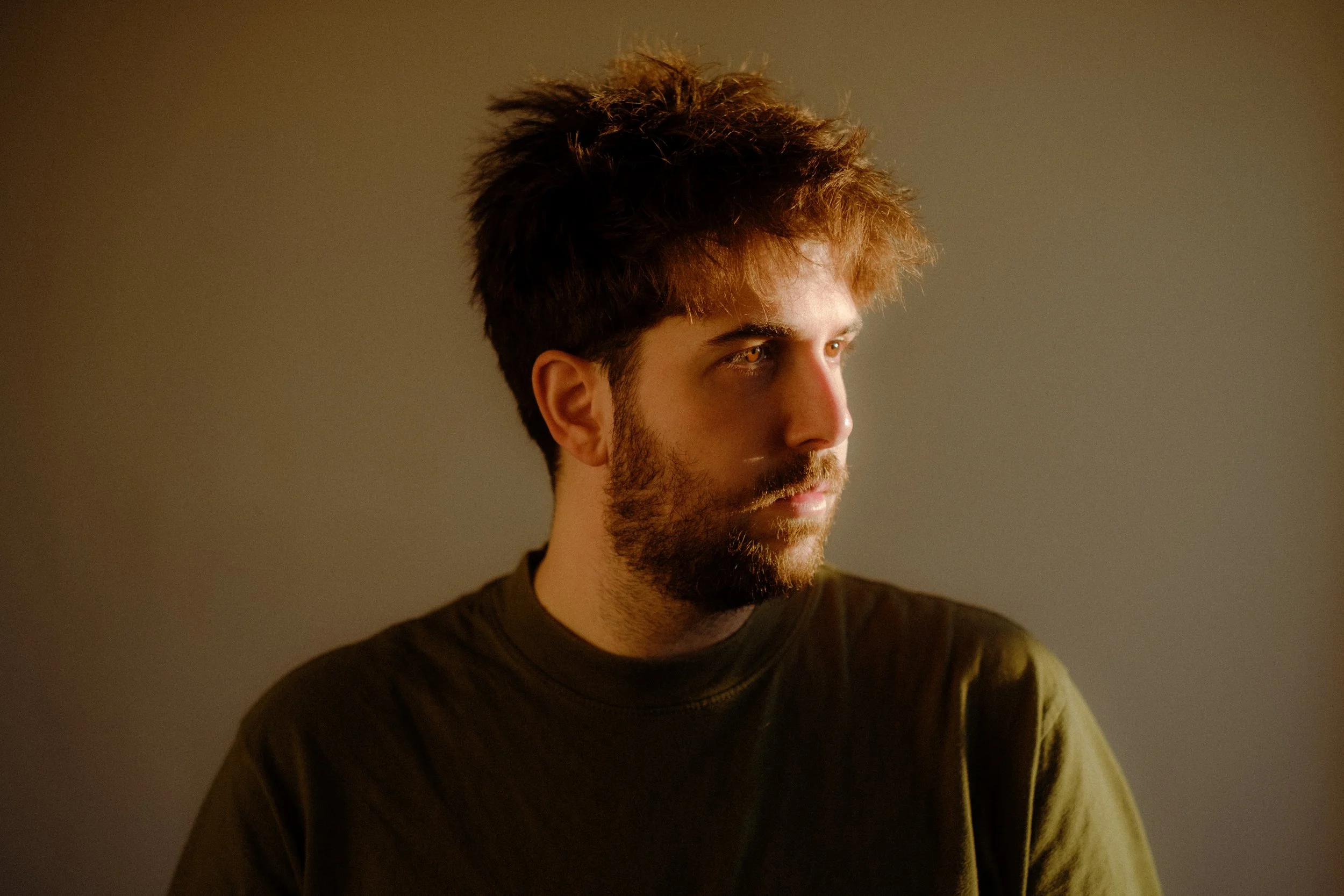 A young man with dark, tousled hair and a beard looking off to the side against a plain background.