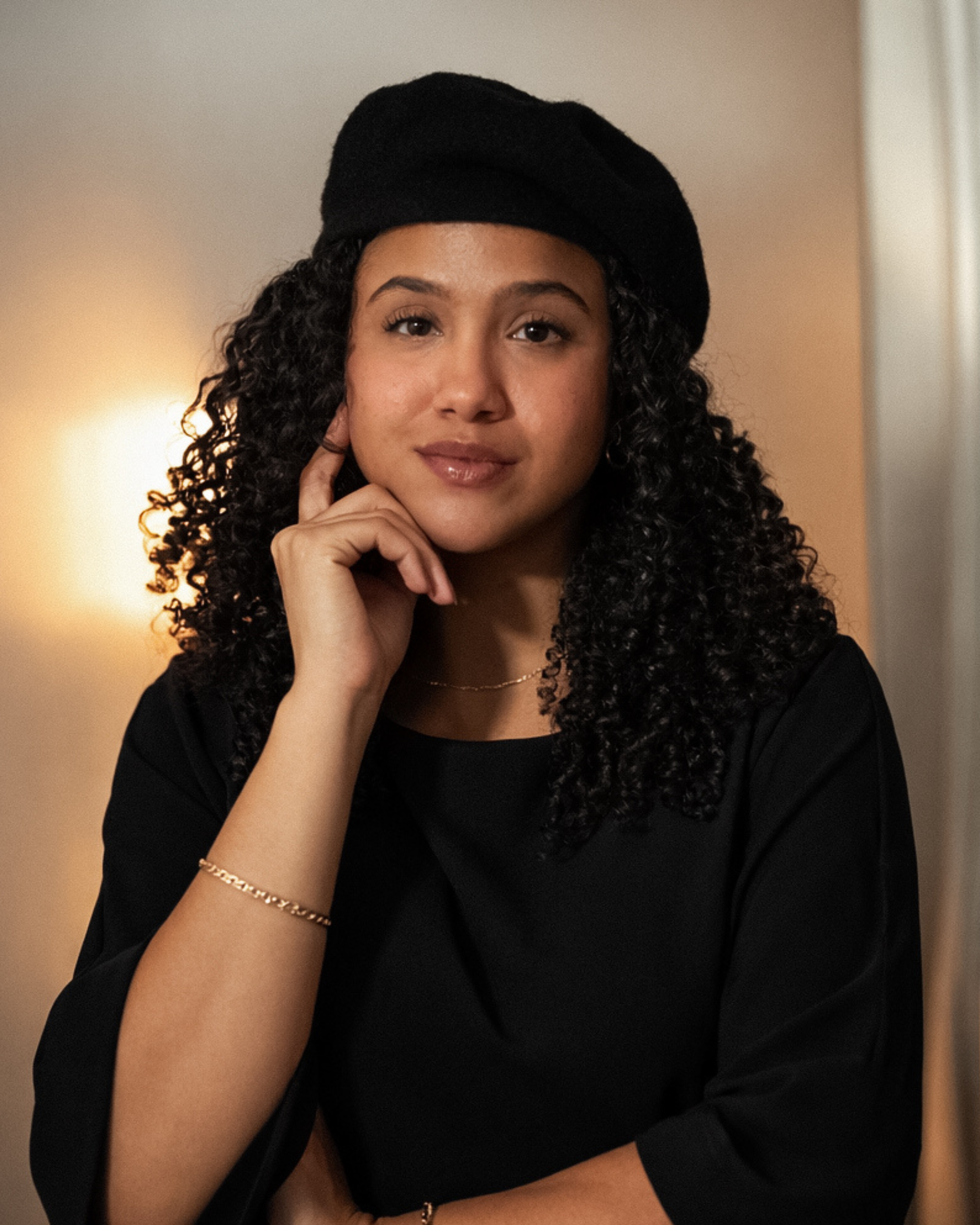 A young woman with curly dark hair wearing a black hat and black top, resting her face on her hand, and looking into the camera with a slight smile.