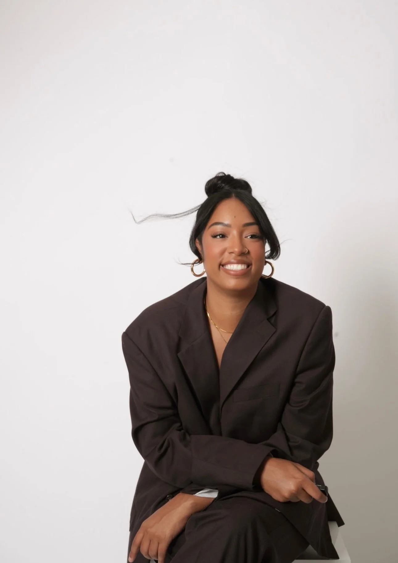Smiling woman in black blazer and gold jewelry sitting against a white background.