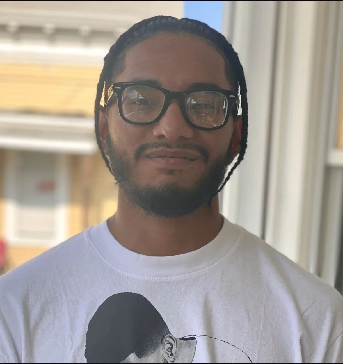 A man with sunglasses, a beard, and dreadlocks, standing indoors near a window, wearing a white T-shirt with a black and white image of a person's head printed on it.