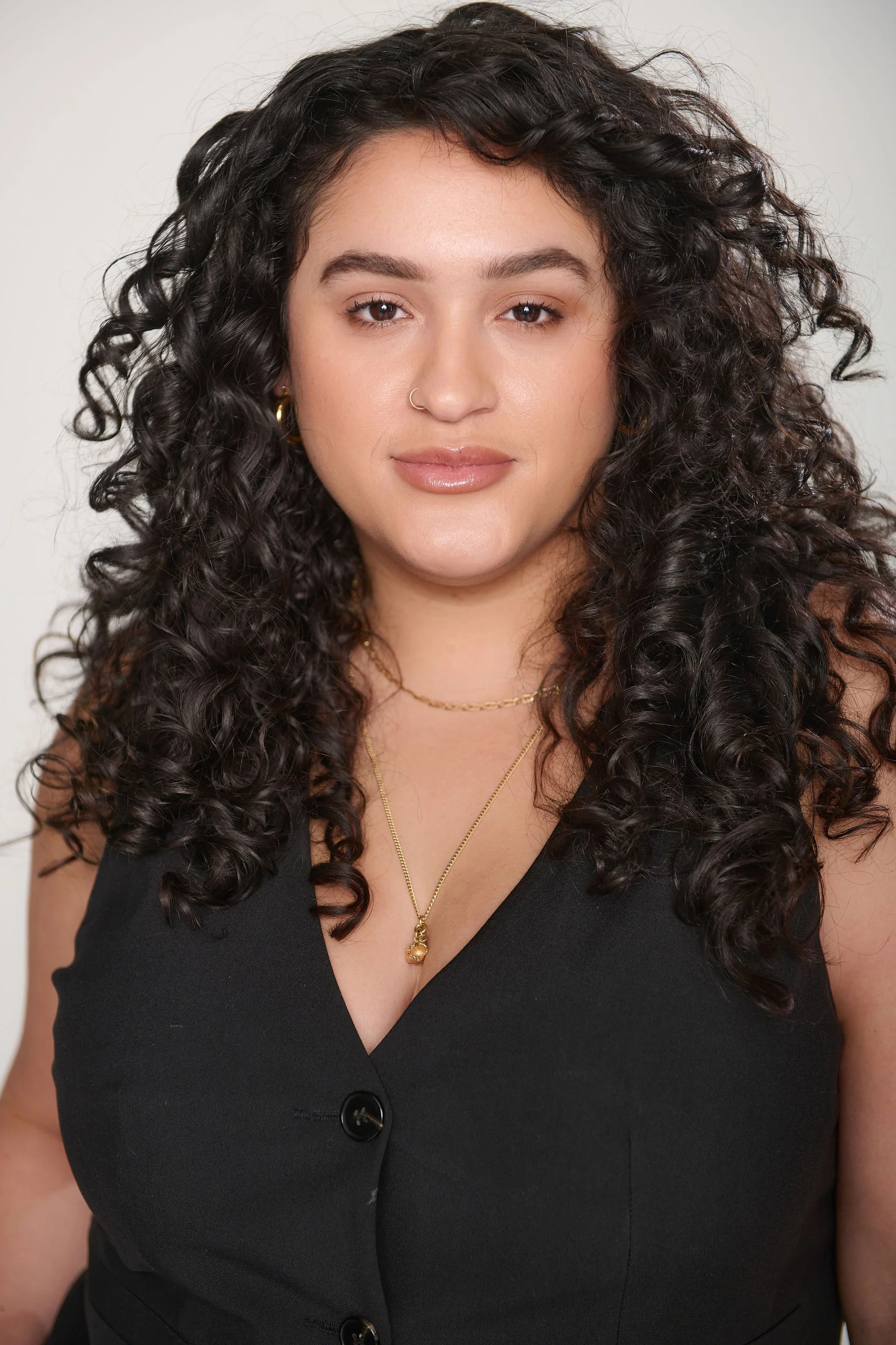 A woman with curly dark hair, wearing a black sleeveless top, gold jewelry, and a nude lipstick, is looking at the camera with a neutral expression.