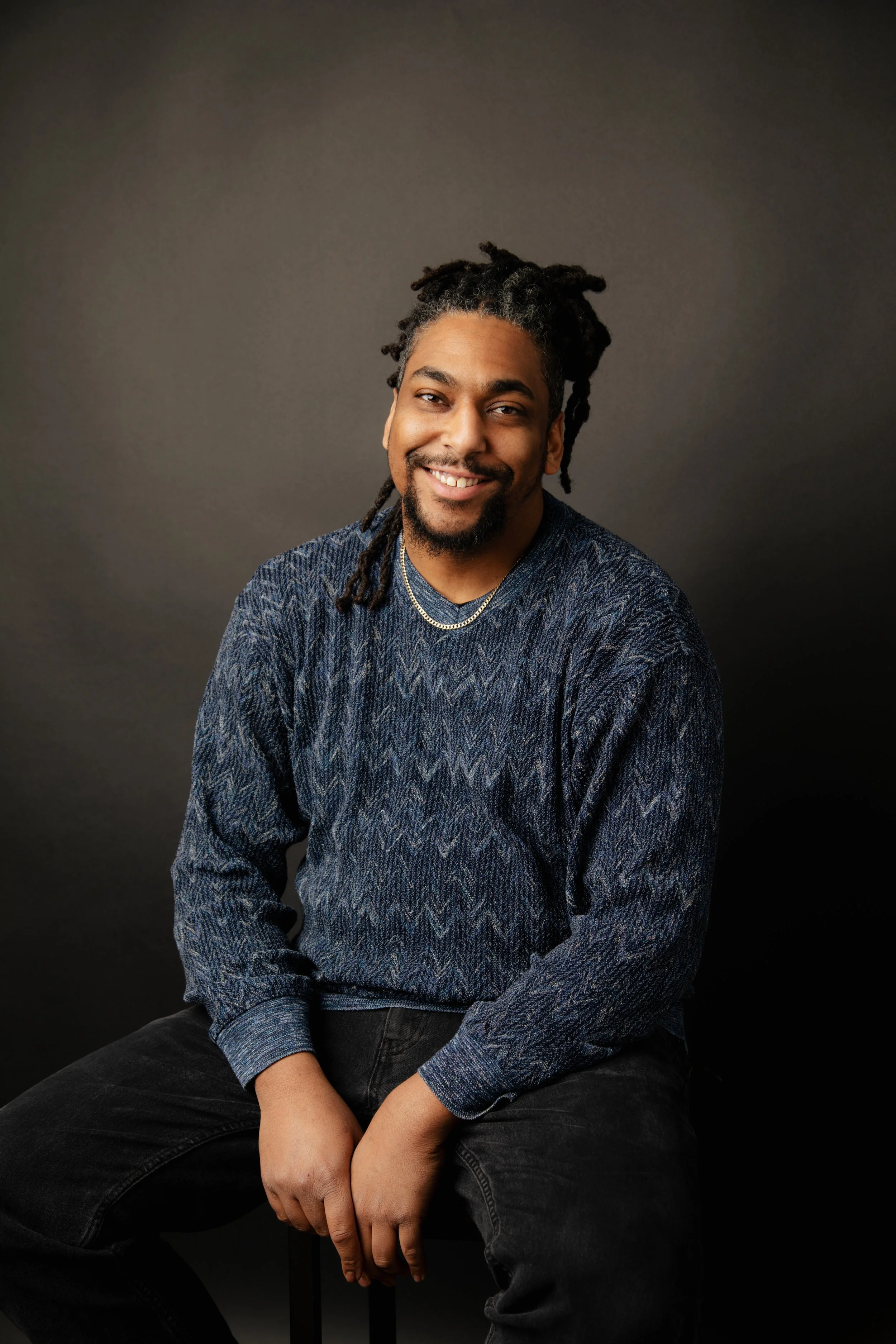 A smiling man with dreadlocks, wearing a patterned blue sweater, black pants, and layered necklaces, sitting against a dark background.