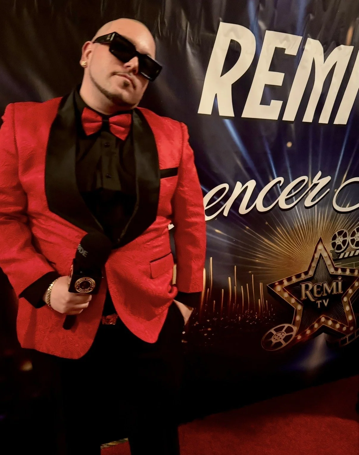 A man dressed in a red tuxedo jacket with black lapels, black shirt, and red bow tie, wearing sunglasses and holding a microphone, standing in front of a backdrop with the words 'Remix concert' and a star with 'Remi TV' written inside.