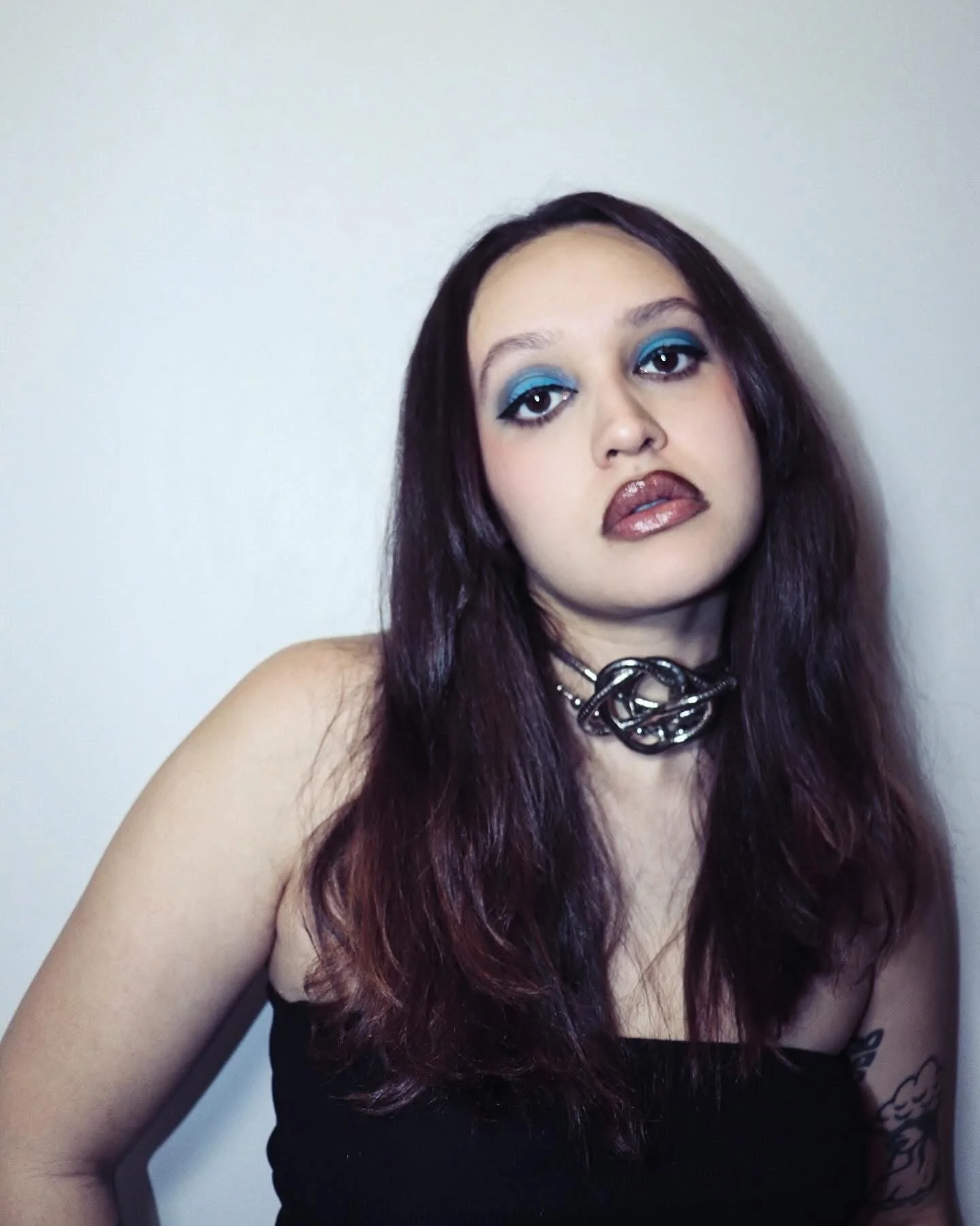 A woman with shoulder-length dark purple hair, wearing bold blue eyeshadow, dark lipstick, a black strapless top, a chunky silver chain necklace, and a tattoo on her left arm, standing against a plain white wall.